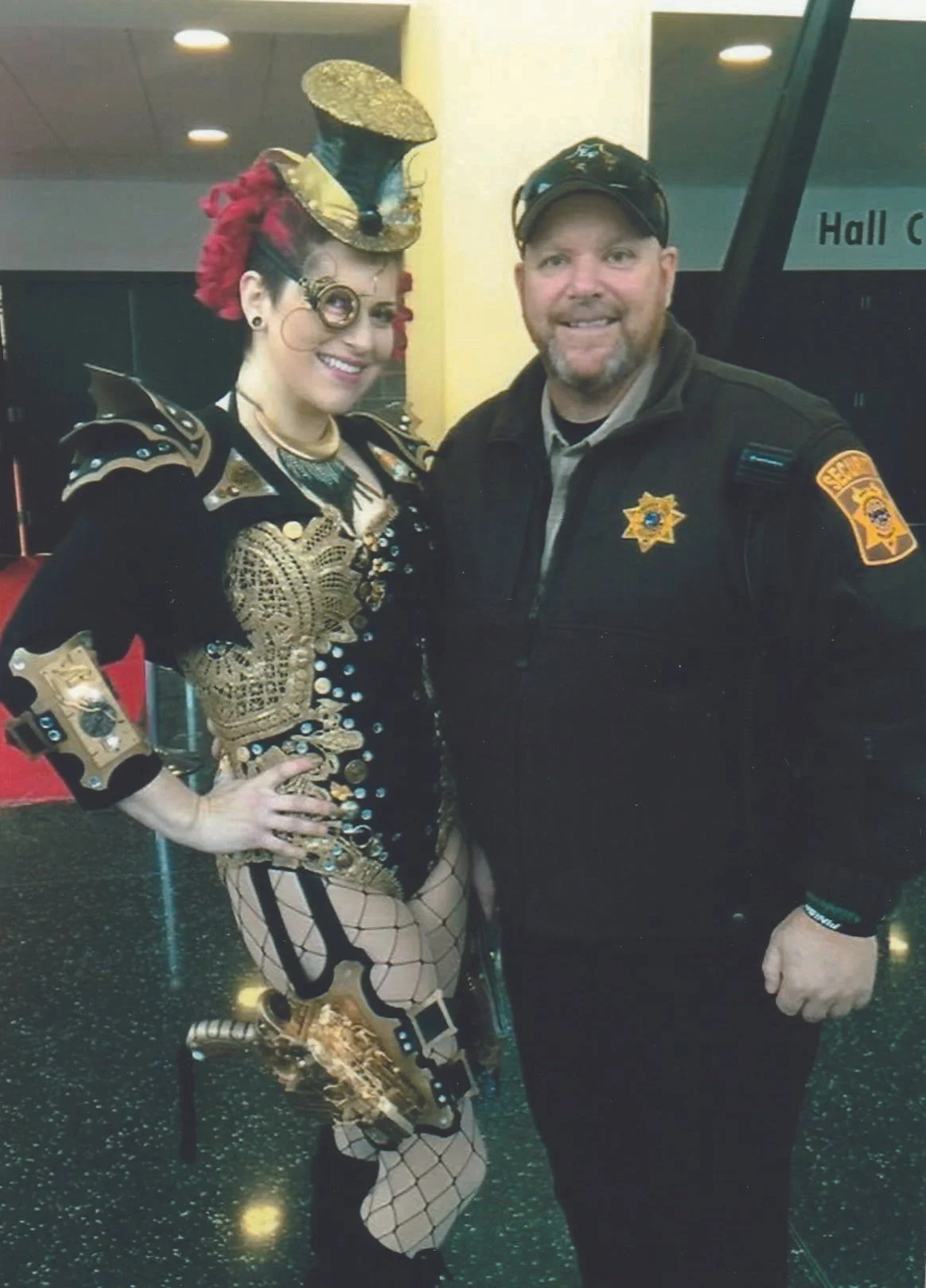 A woman dressed in a steampunk costume with a top hat and fishnet stockings, standing next to a man in a sheriff uniform, both smiling indoors.
