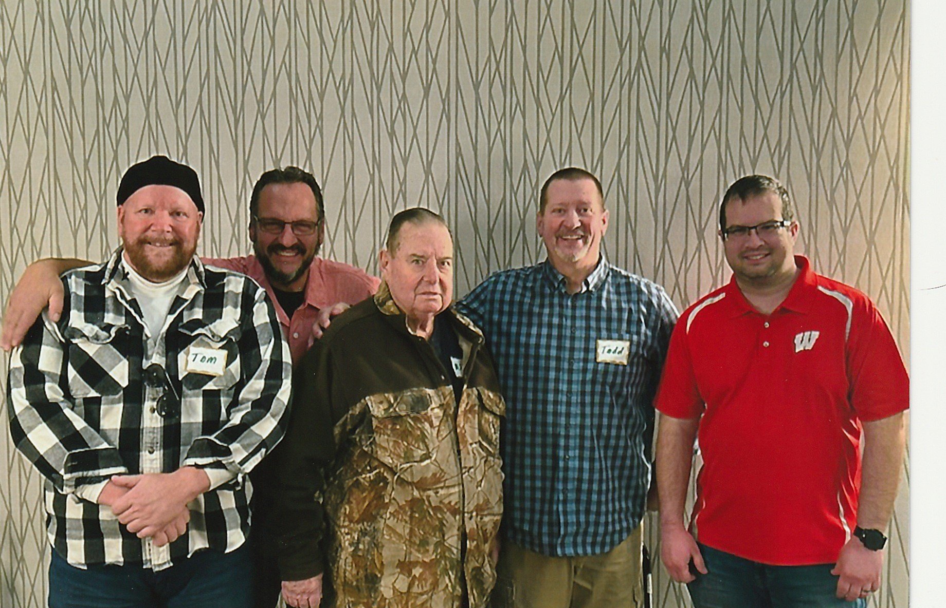 Group of five men standing together indoors in front of a patterned wall, smiling at the camera, some wearing glasses, two with name tags, one wearing a camo jacket, with varied casual clothing.