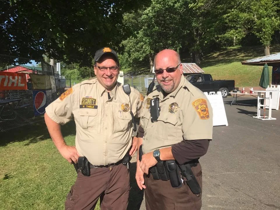 Two male security officers standing outdoors, smiling, wearing tan uniforms with security patches, in front of trees, a fence, and parked vehicles.