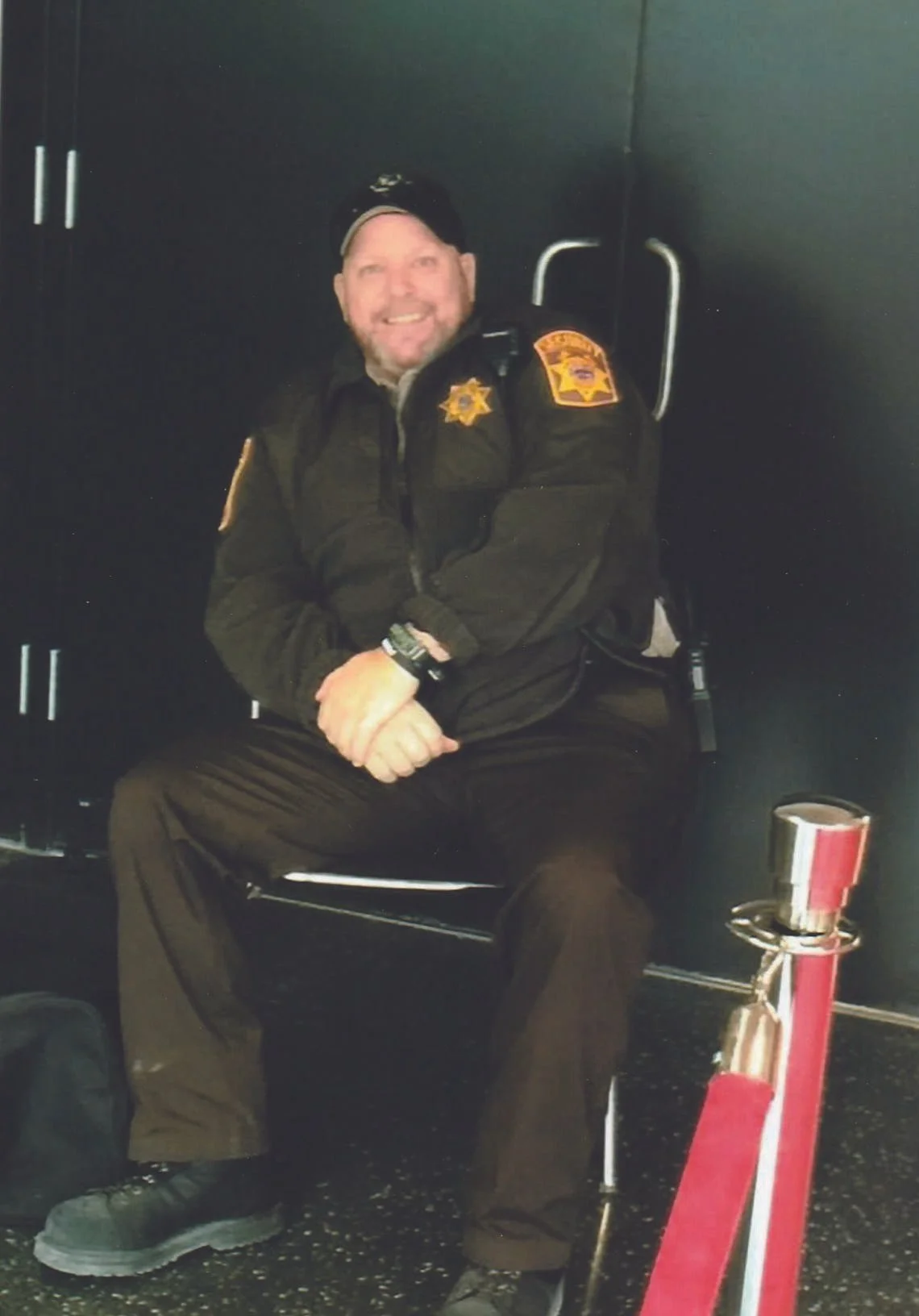 Man in police uniform sitting on a bench, smiling, with a black wall in the background and a red and silver stanchion nearby.