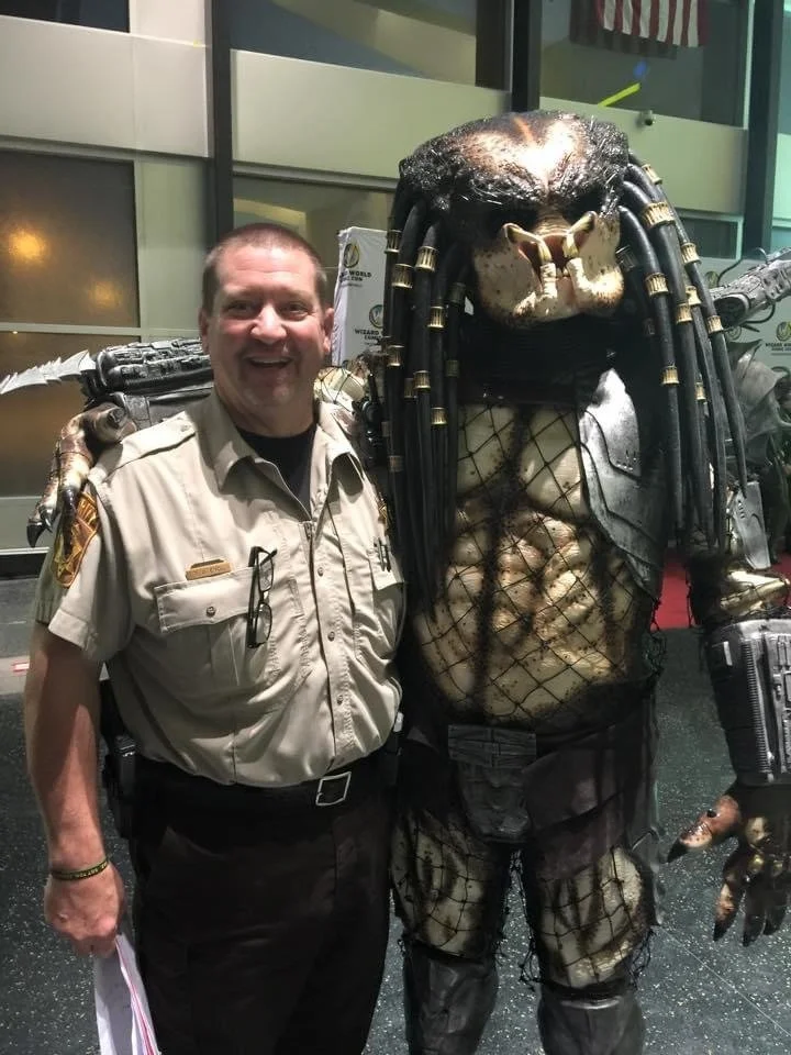 A man in a sheriff's uniform standing next to a large Predator costume, both smiling.