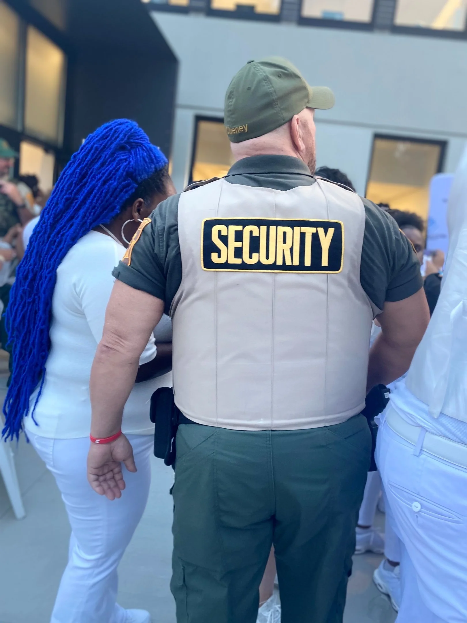 Security personnel standing at an event, viewed from behind, wearing a vest with 'SECURITY' written on it and a cap, next to a woman with blue dreadlocks and white attire.