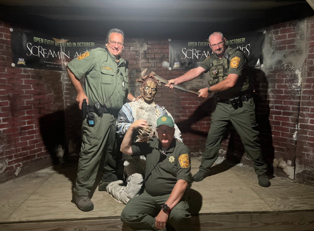 Three men dressed as park rangers or law enforcement officers posing with a Halloween zombie prop in a brick wall setting, with a skeleton design. One man is sitting on the floor flexing his arm, another man is standing and holding a large knife, and
