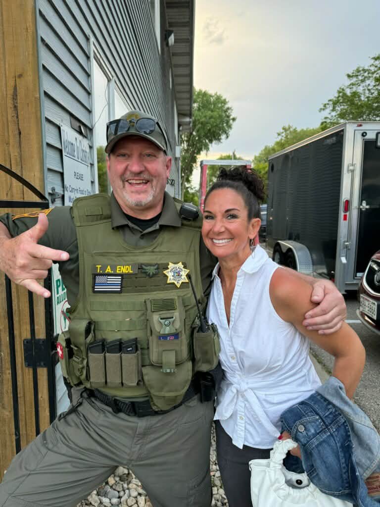 A police officer and a woman smiling and posing together outdoors, with police trucks in the background.