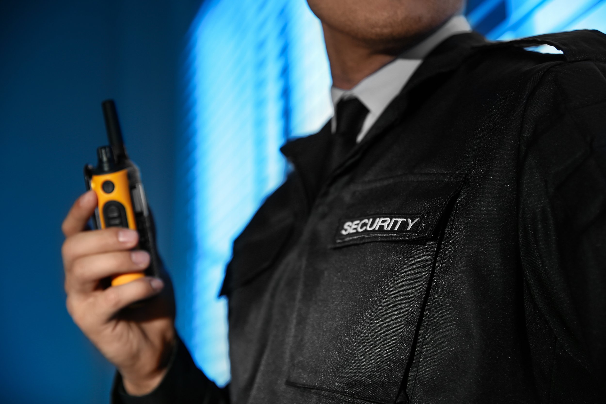 Close-up of security guard wearing black uniform with 'SECURITY' patch, holding a walkie-talkie, against a blue background.