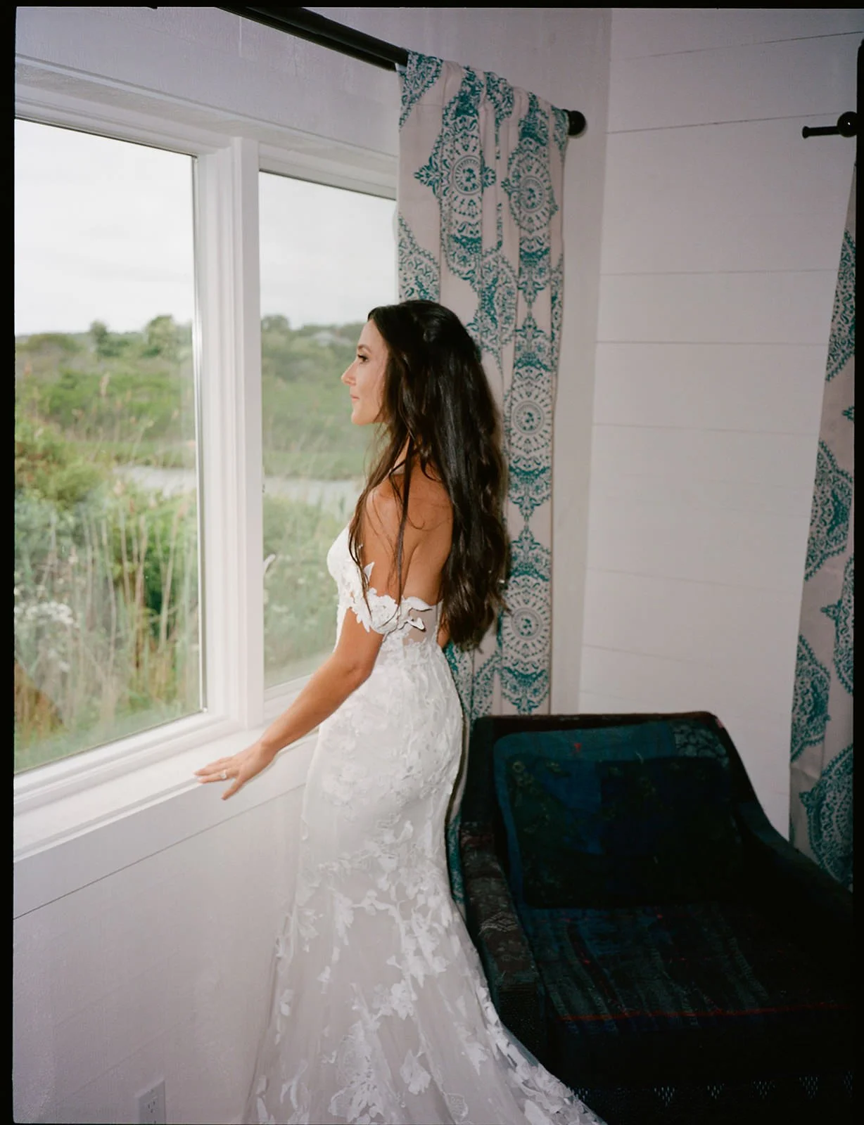 Bride in a lace gown looking out the window at the marsh before her Crow's Nest wedding in Montauk, shot on film