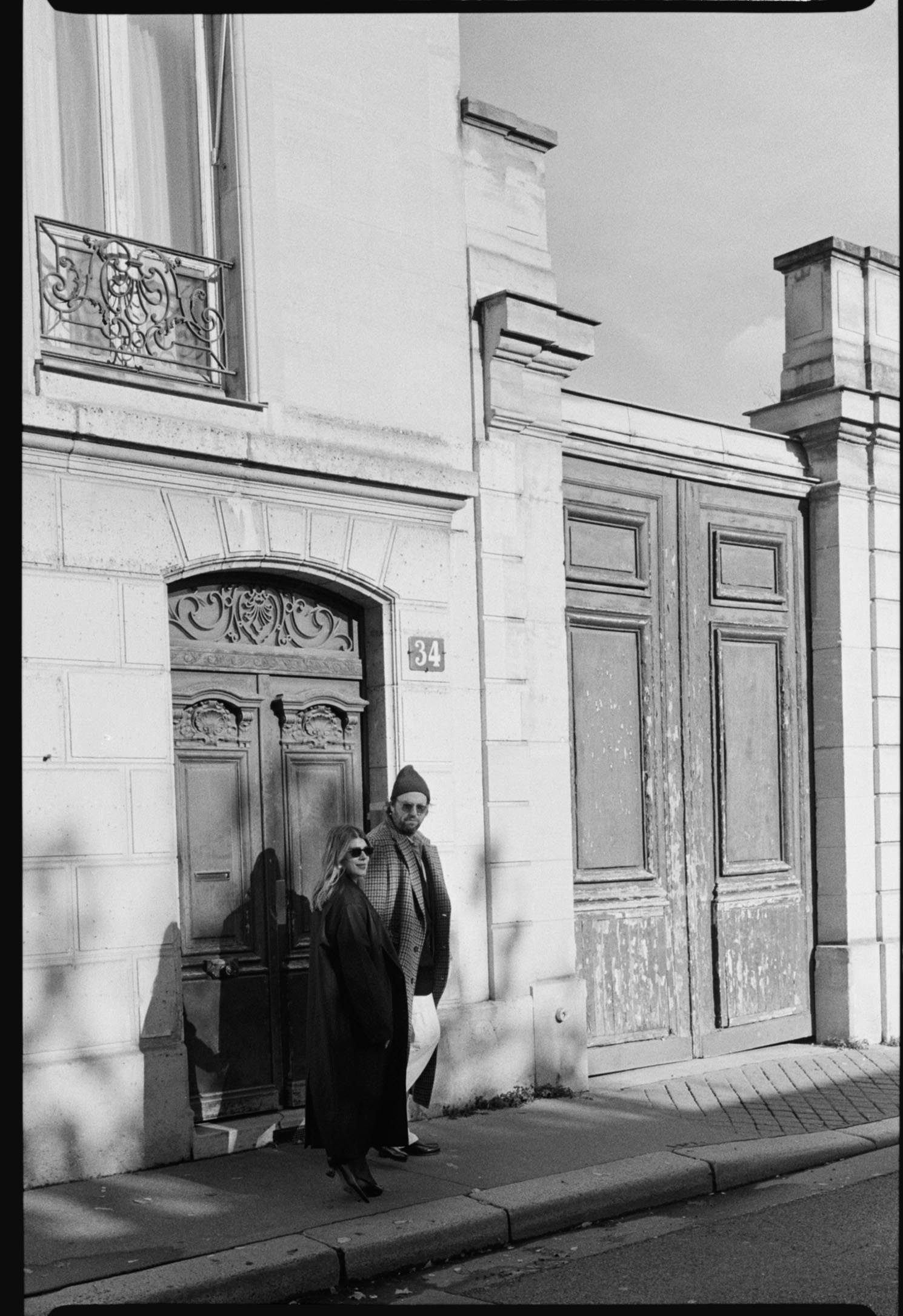A black and white photo of a man and woman standing outside on a city sidewalk in front of a building with ornate wooden doors and window. The man is wearing glasses, a beanie, and a checked coat, while the woman is wearing sunglasses and a long dark coat.
