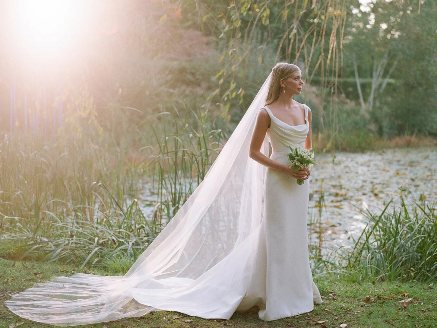 Bride standing outdoors by a pond, holding a bouquet, in a white wedding dress and veil, with sunlight filtering through trees.