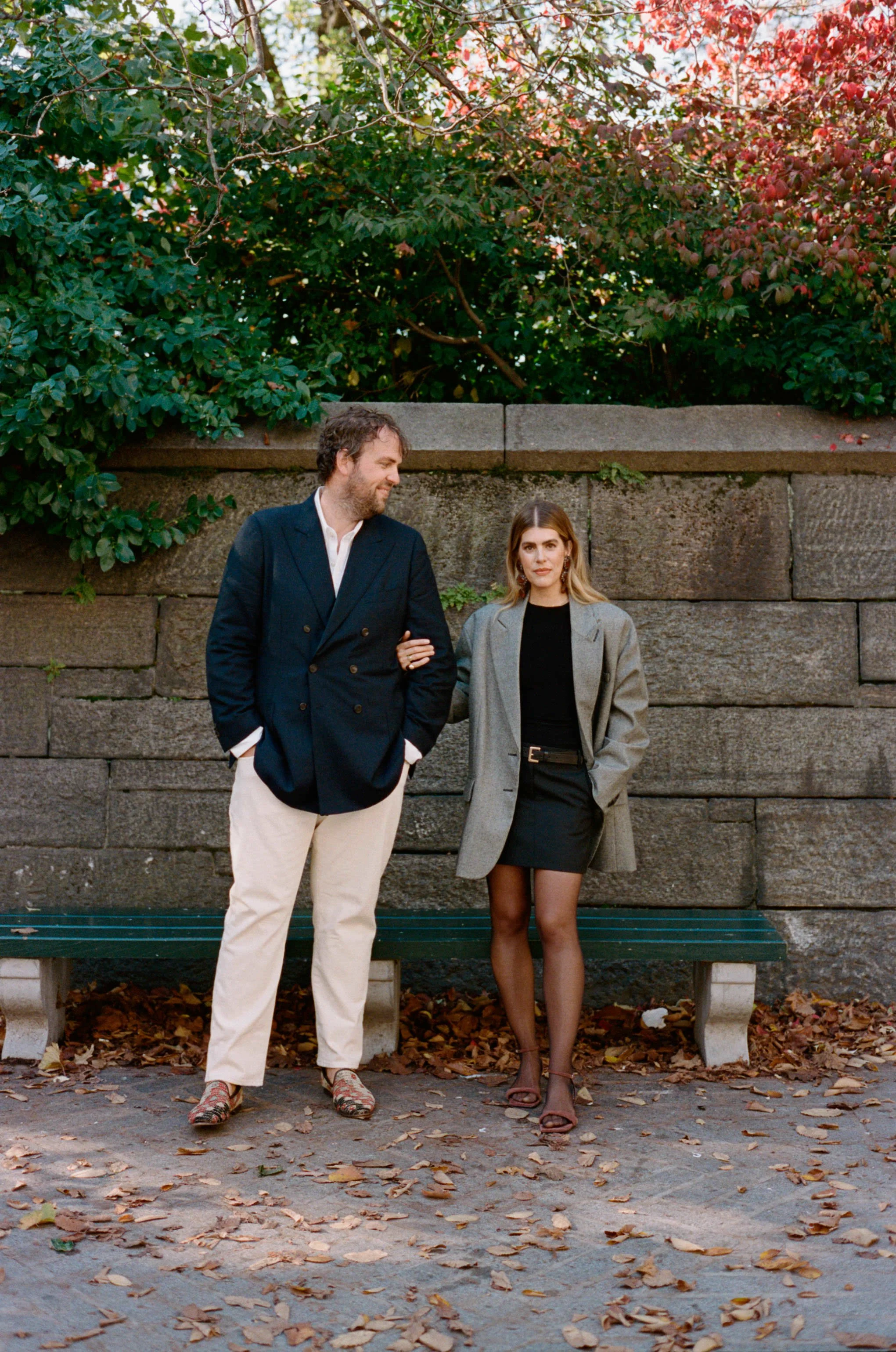 A man and woman walking arm in arm outdoors during fall, with leaves on the ground and a stone wall behind them. The man wears a dark blazer and white pants; the woman wears a gray coat over a black outfit.