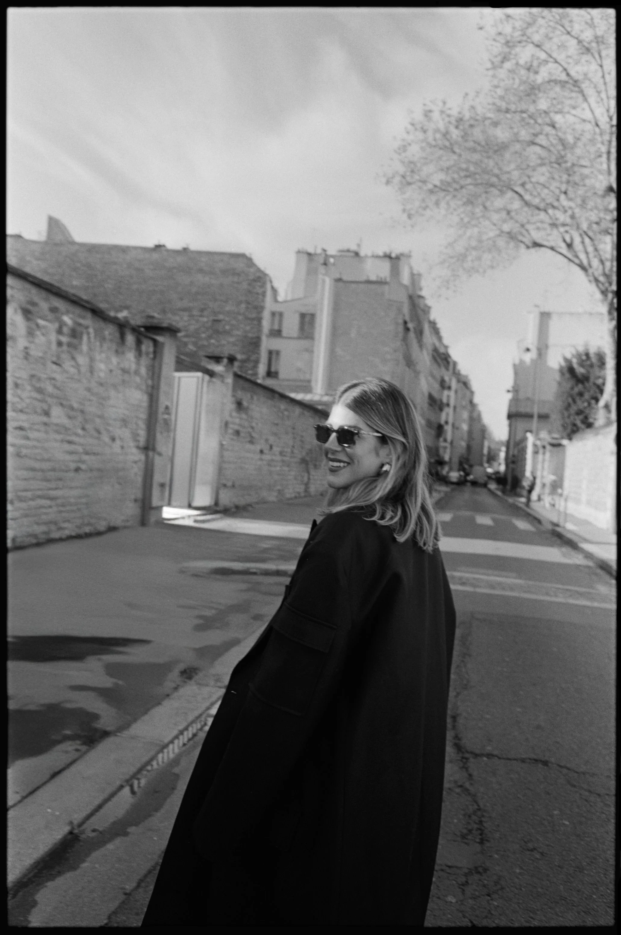 A woman with shoulder-length hair wearing sunglasses and a dark coat standing on a city street, smiling and looking over her shoulder.