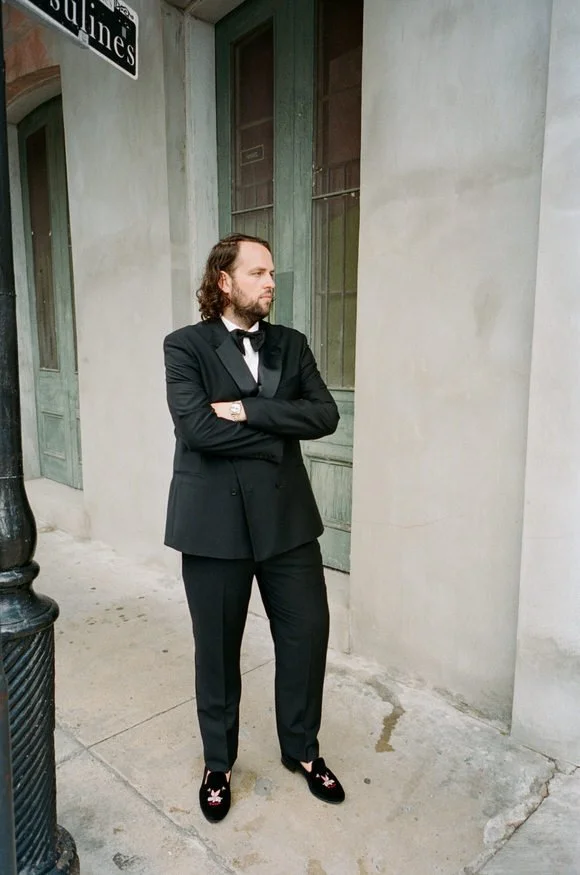 Man in a black tuxedo standing with arms crossed outside a building with green doors.