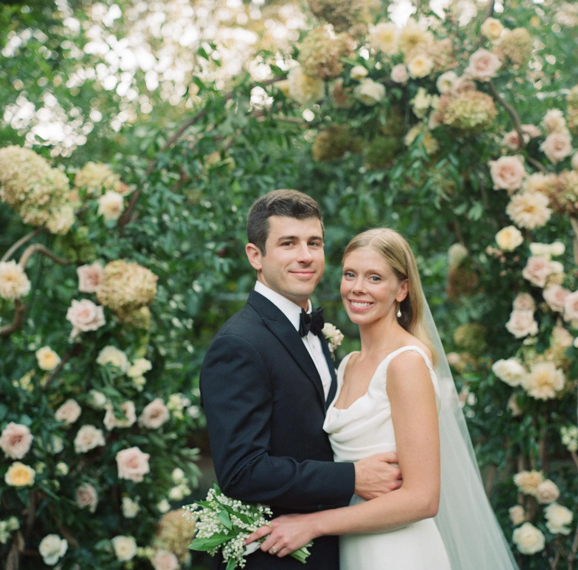 Bride and groom smiling in front of a wall of blush garden roses in East Hampton