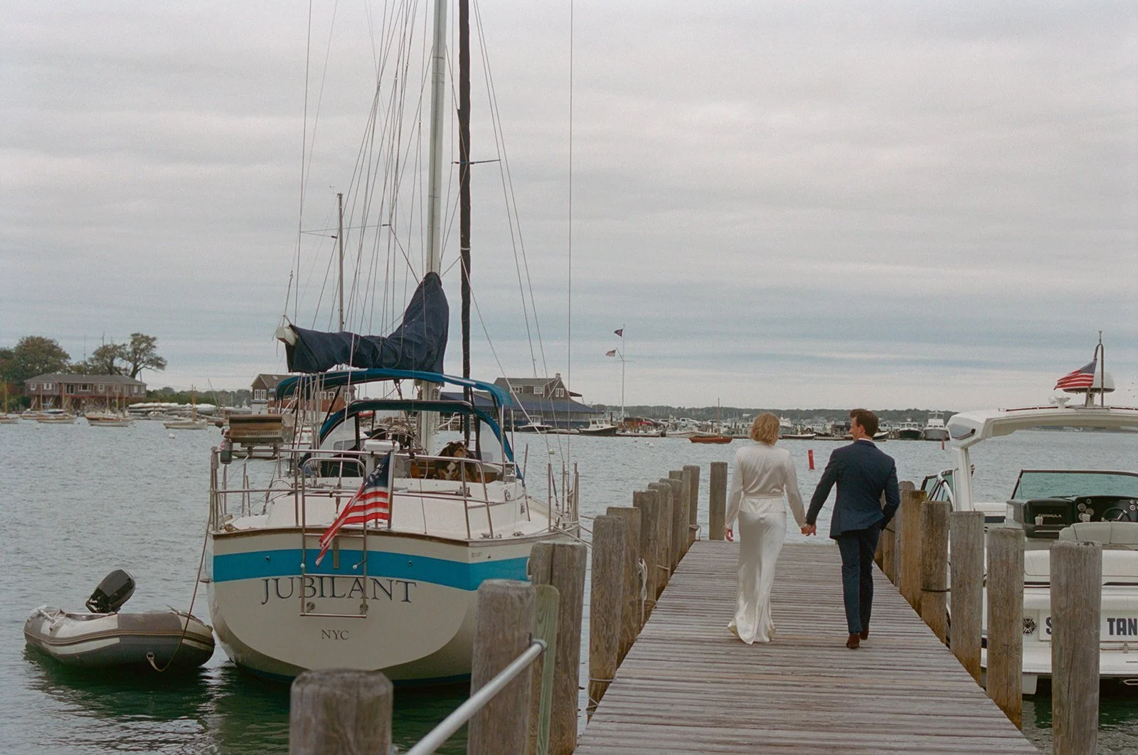 Bride and groom walking hand in hand down a wooden dock past a moored sailboat on Shelter Island