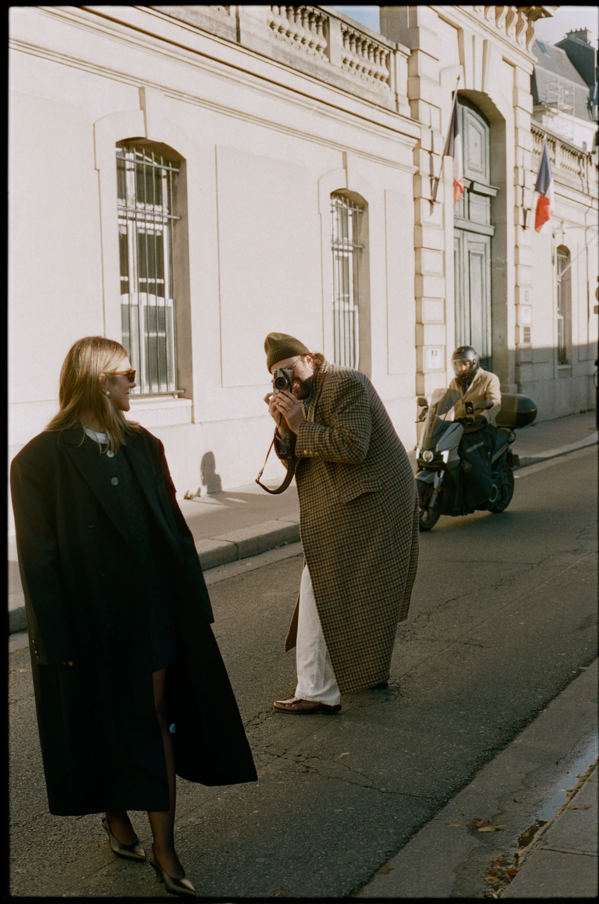 A woman with sunglasses and a coat standing on a city street being photographed by a man holding a camera. A person on a scooter is passing by in the background near a historic building with French flags.