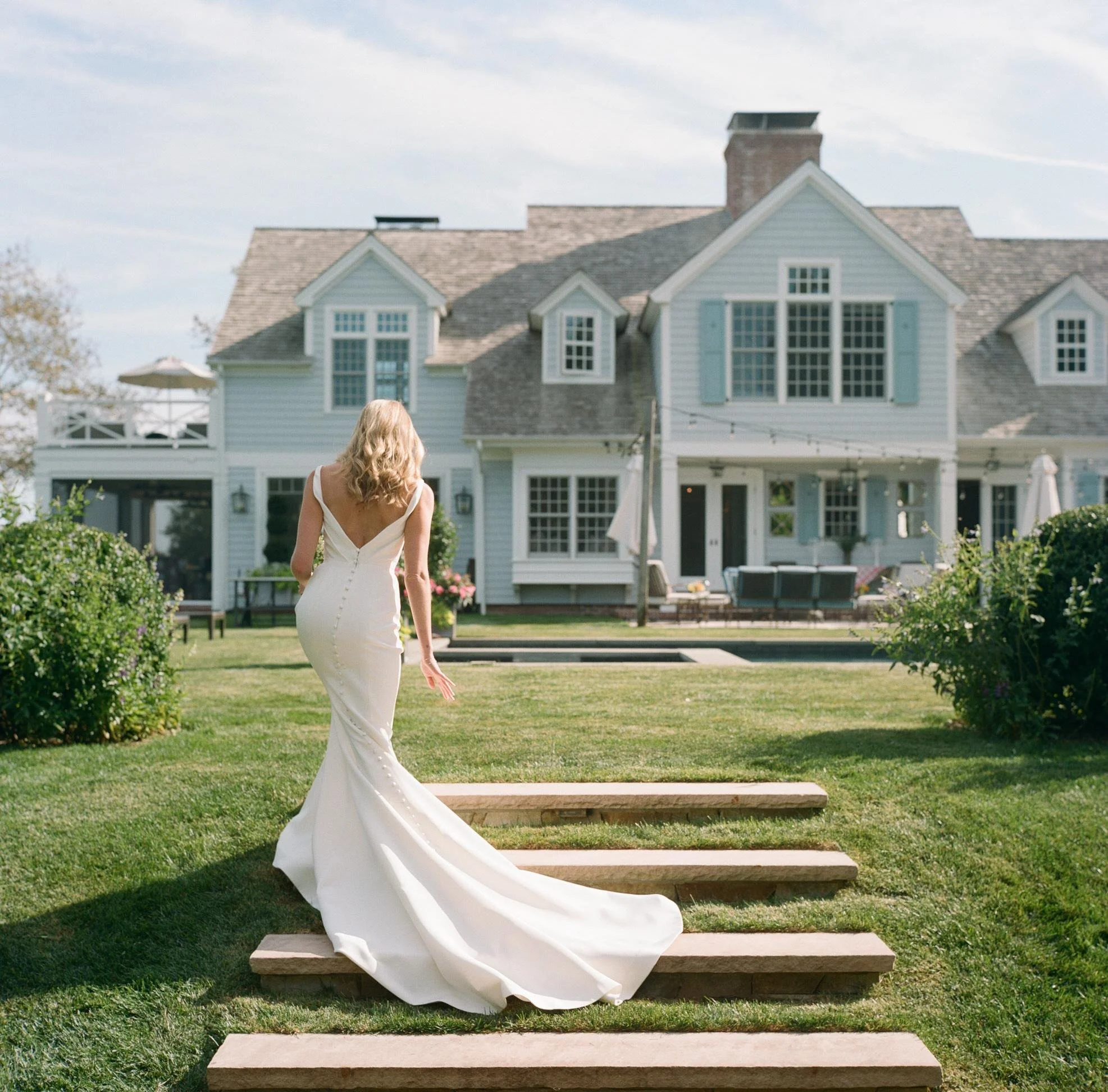 Bride walking up the lawn steps toward a gray shingled house in East Hampton