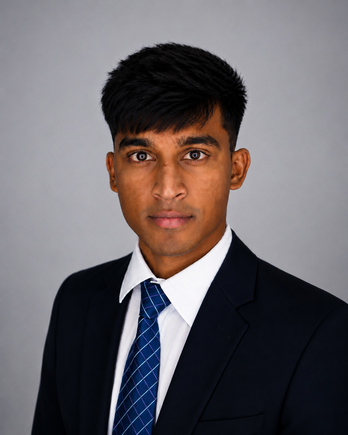 A young man with dark hair and brown skin wearing a black suit, white shirt, and blue plaid tie, looking directly at the camera against a plain gray background.