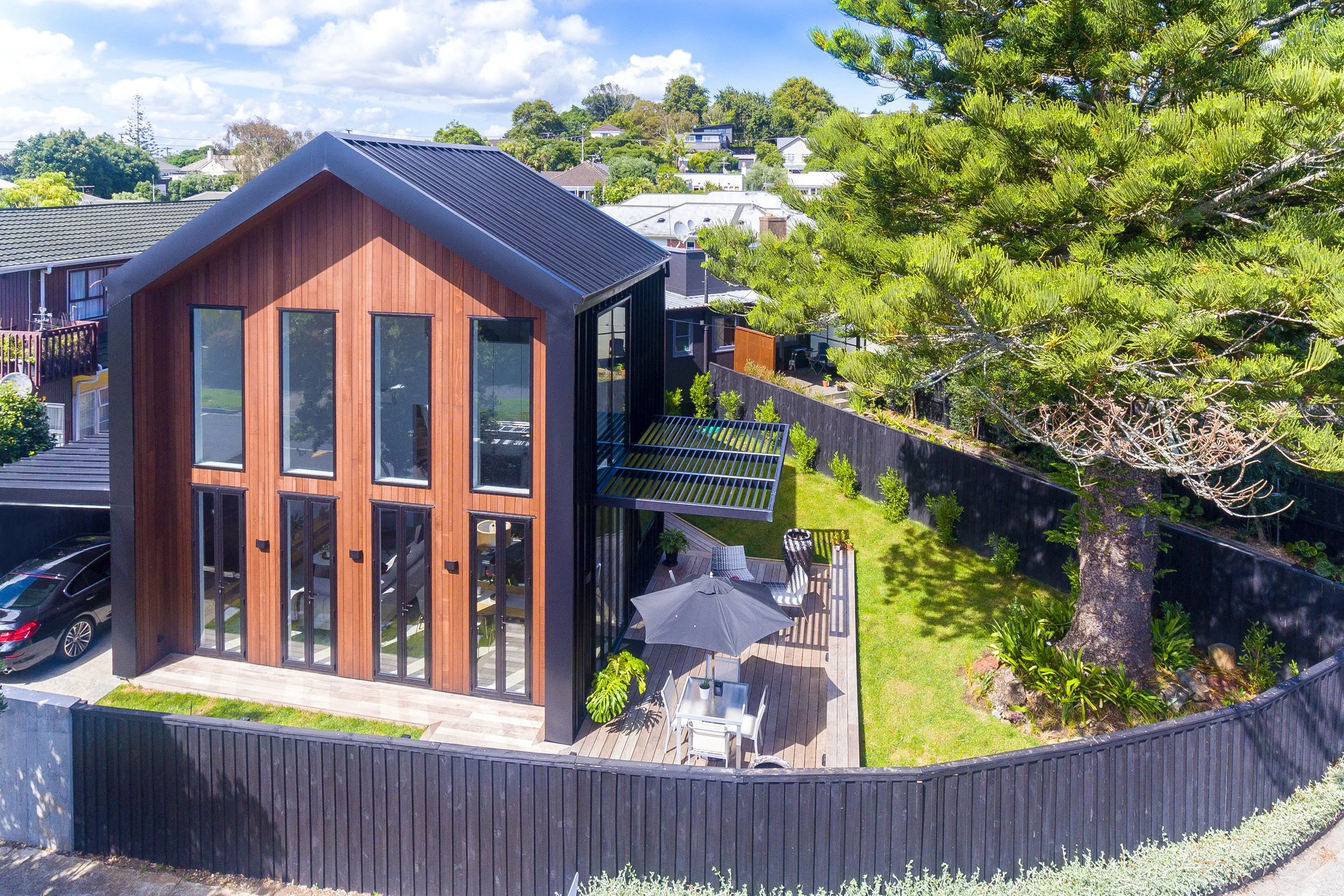 A modern two-story house with a wooden and black exterior, large windows, and a backyard patio with outdoor furniture, an umbrella, and lush greenery.