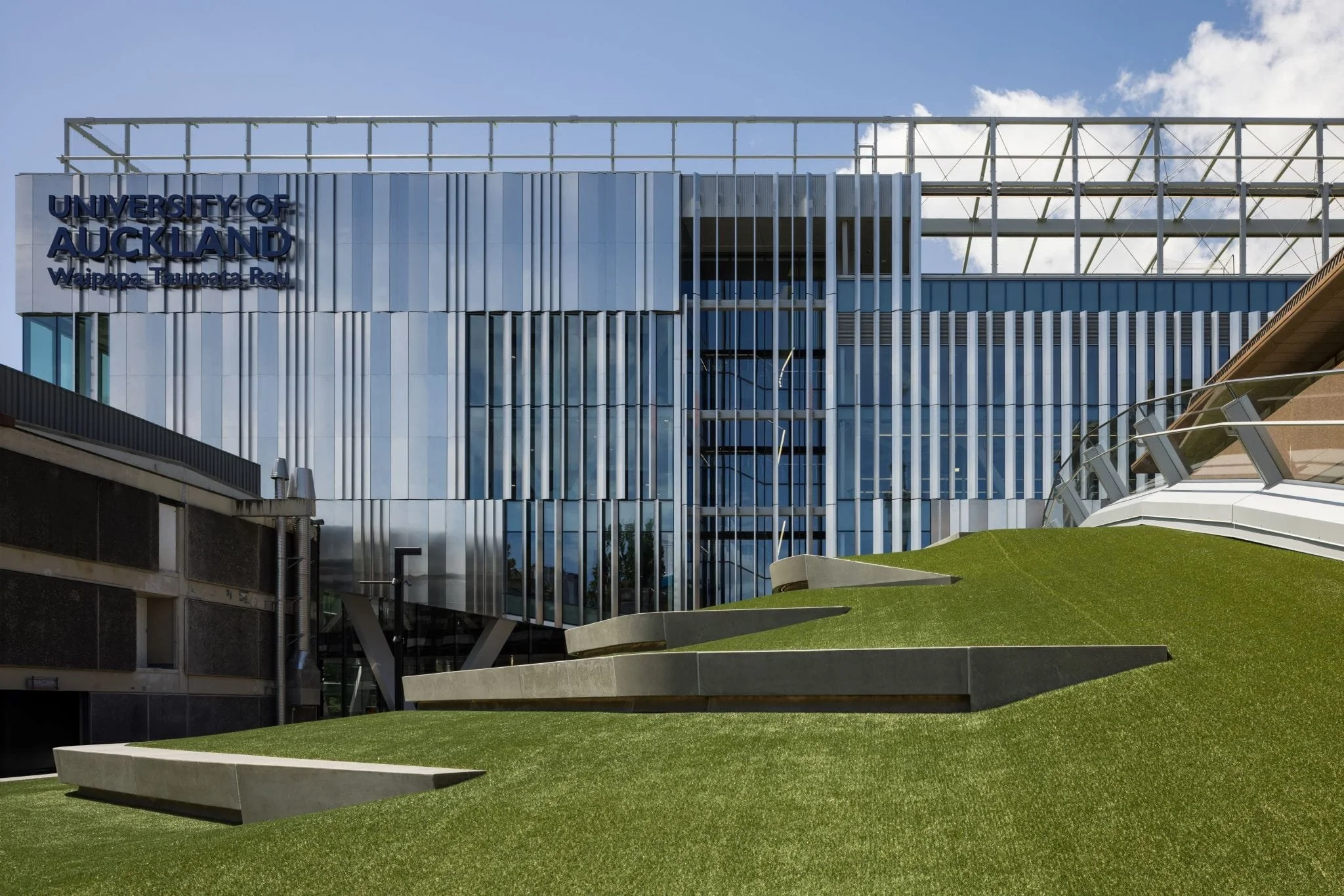 Exterior view of the University of Auckland building with modern glass architecture and a green artificial grass roof in the foreground.