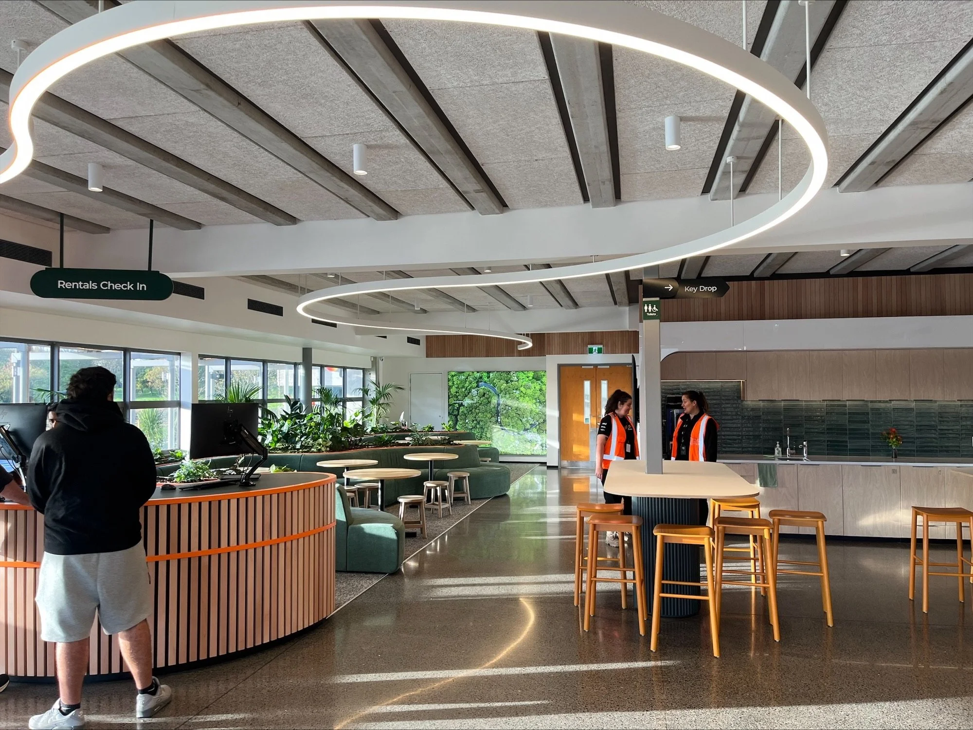 Modern hotel lobby with check-in counter, seating area with green furniture and large windows, and two staff members in orange vests behind a white high table, with a man in a black hoodie at the counter.