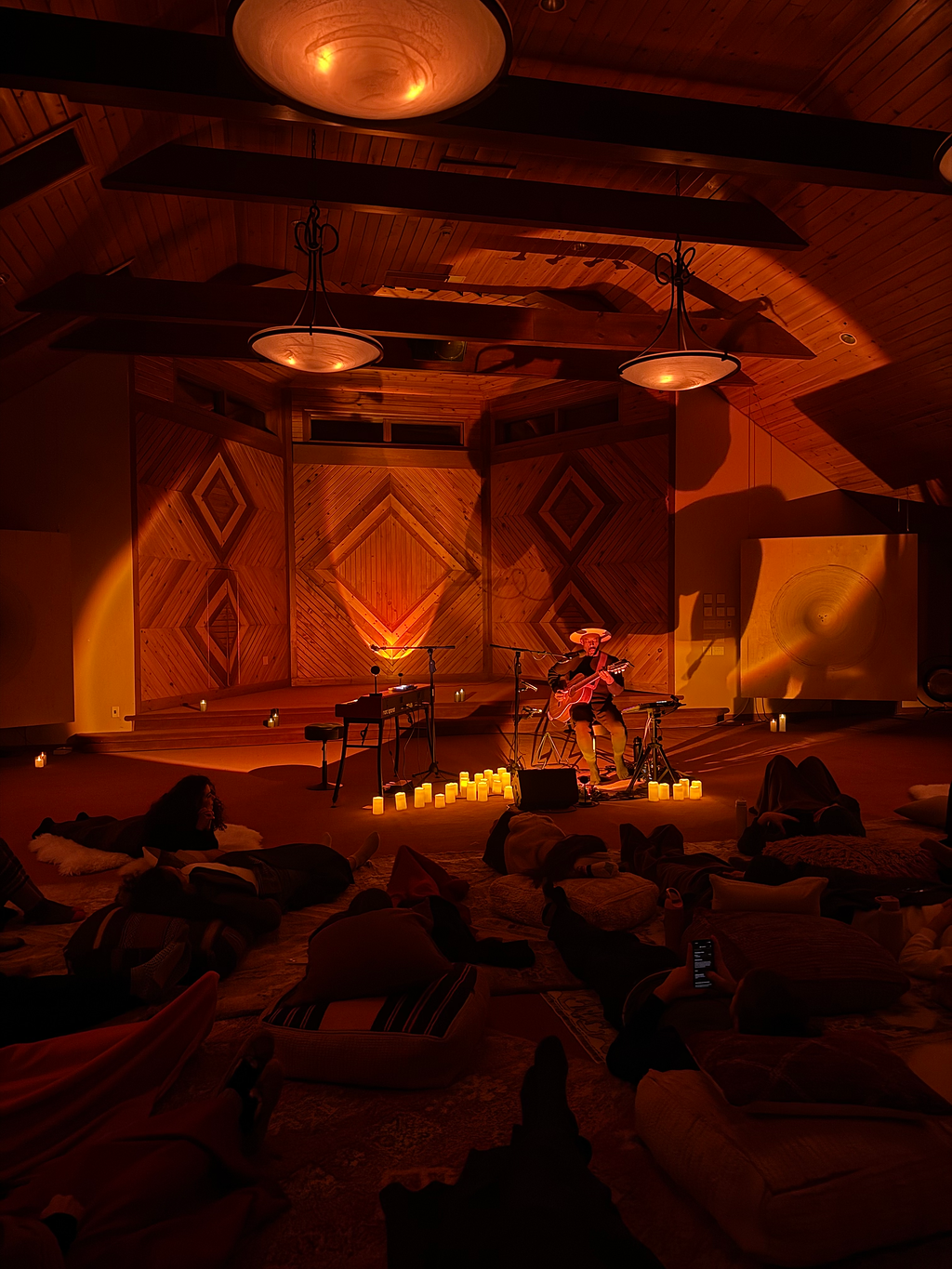 People dressed in white sitting in a circle around a central structure with candles, in an outdoor setting at night with trees and candle lights in the background.