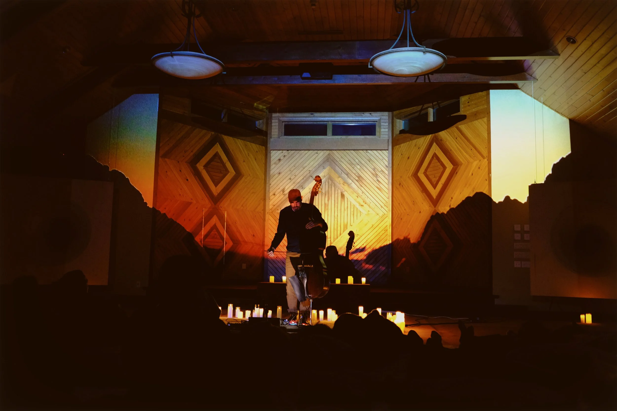 People dressed in white sitting in a circle around a central structure with candles, in an outdoor setting at night with trees and candle lights in the background.