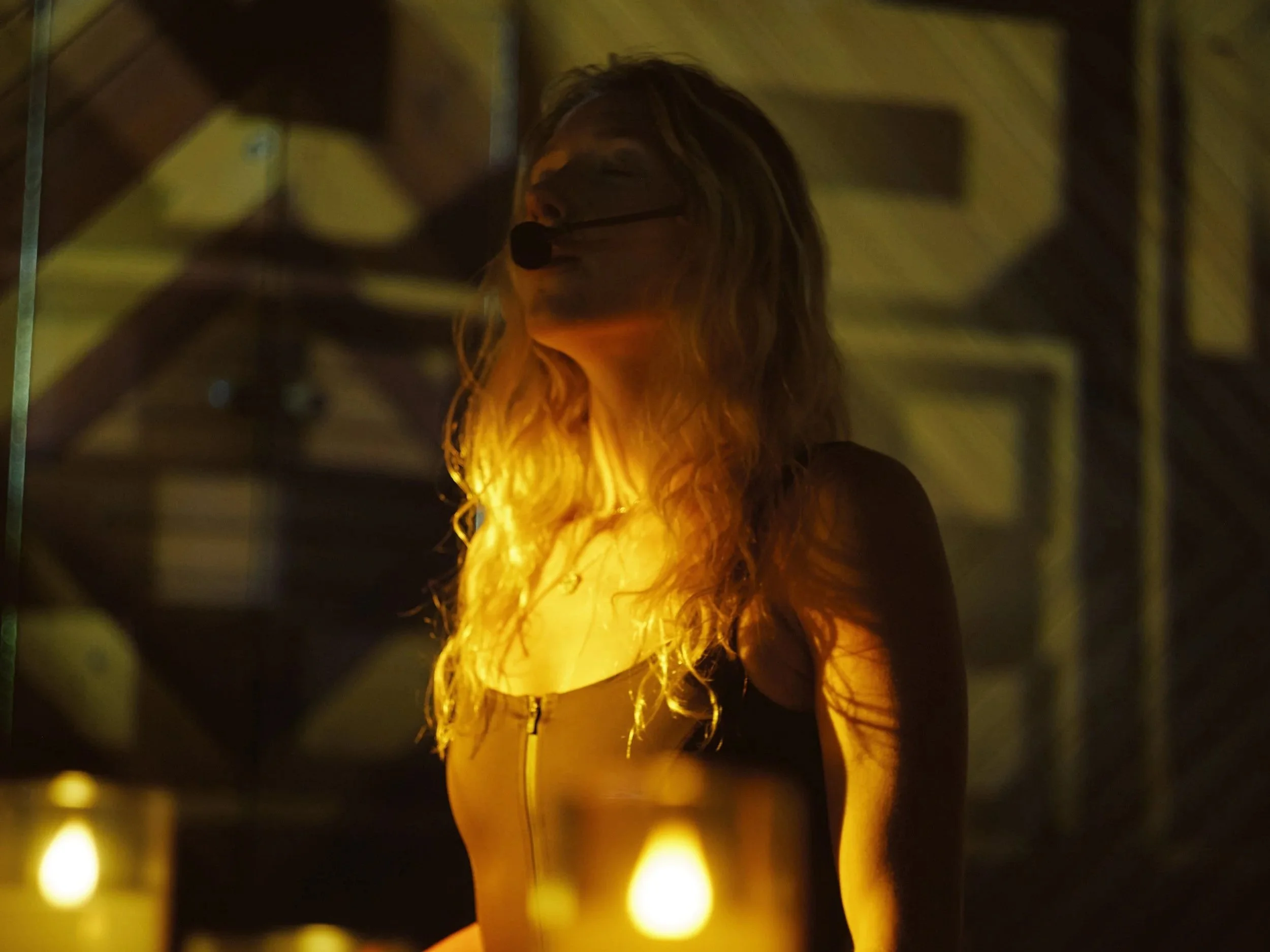 A young woman with long blonde hair sitting on a brown bench in front of an abstract wall installation with draped textured fabric and hanging ribbon-like elements, illuminated by warm lighting.