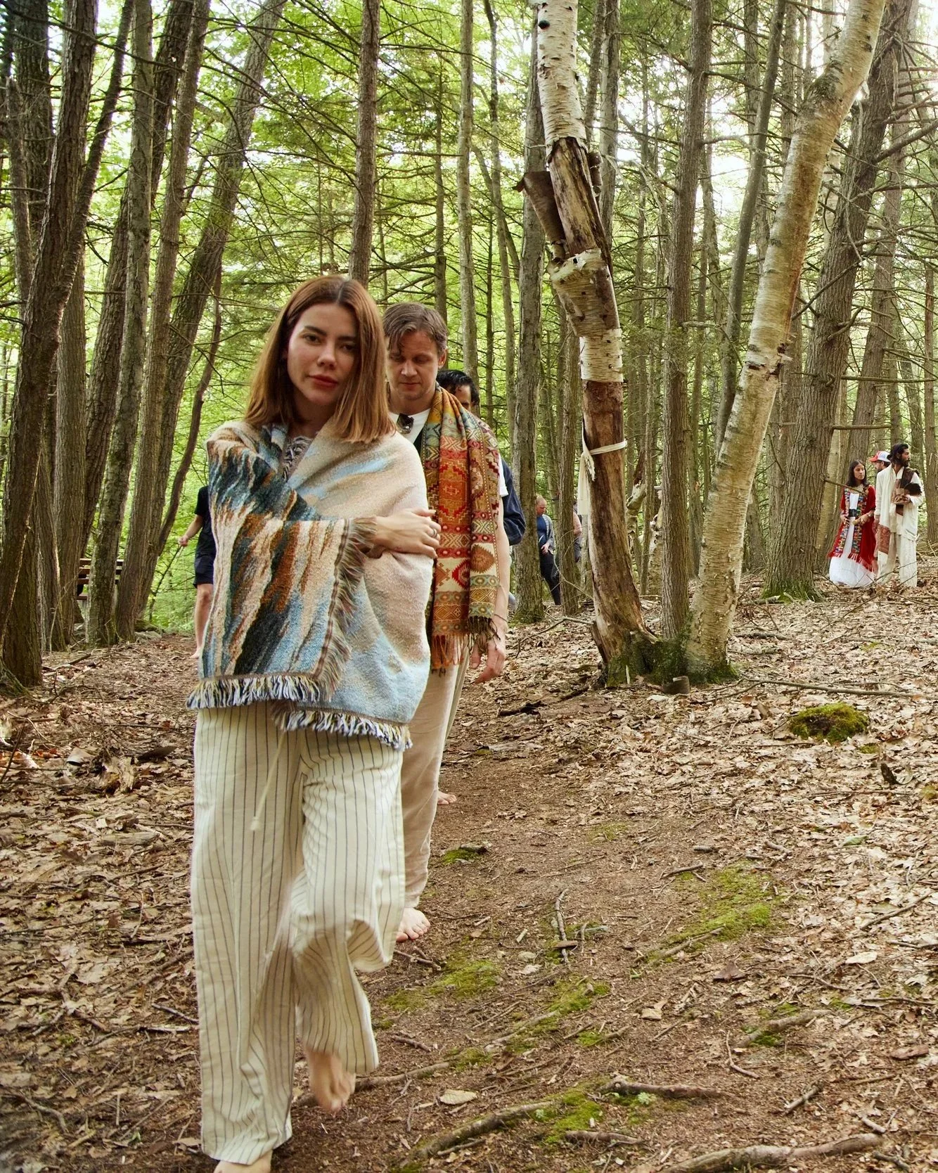 Group of people walking on a trail in a dense forest, some wrapped in blankets or shawls, dressed casually and barefoot, with trees and greenery surrounding them.