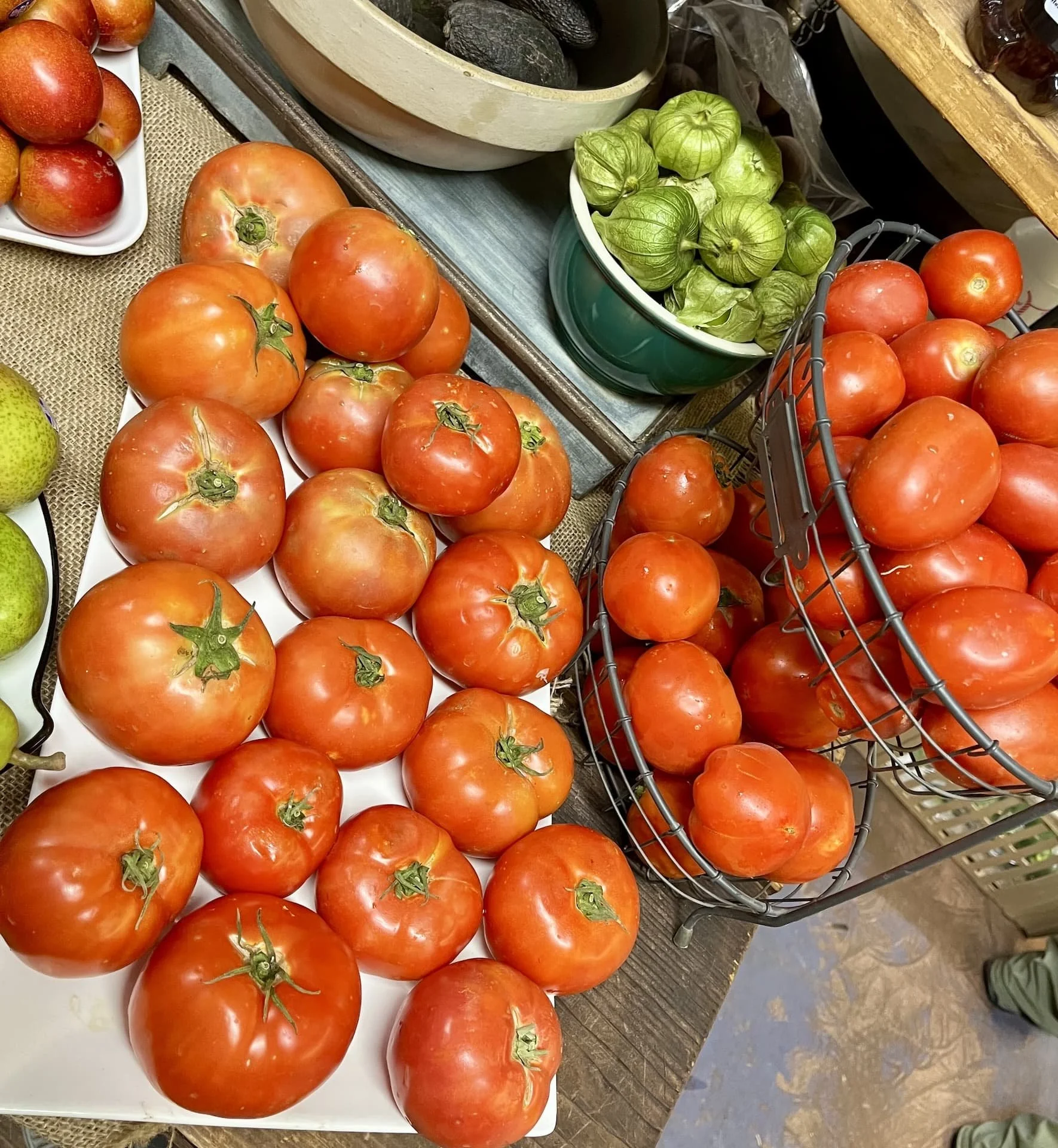 Tomatoes on a plate and in a basket, with green tomatillos in a bowl, and avocados in another bowl.