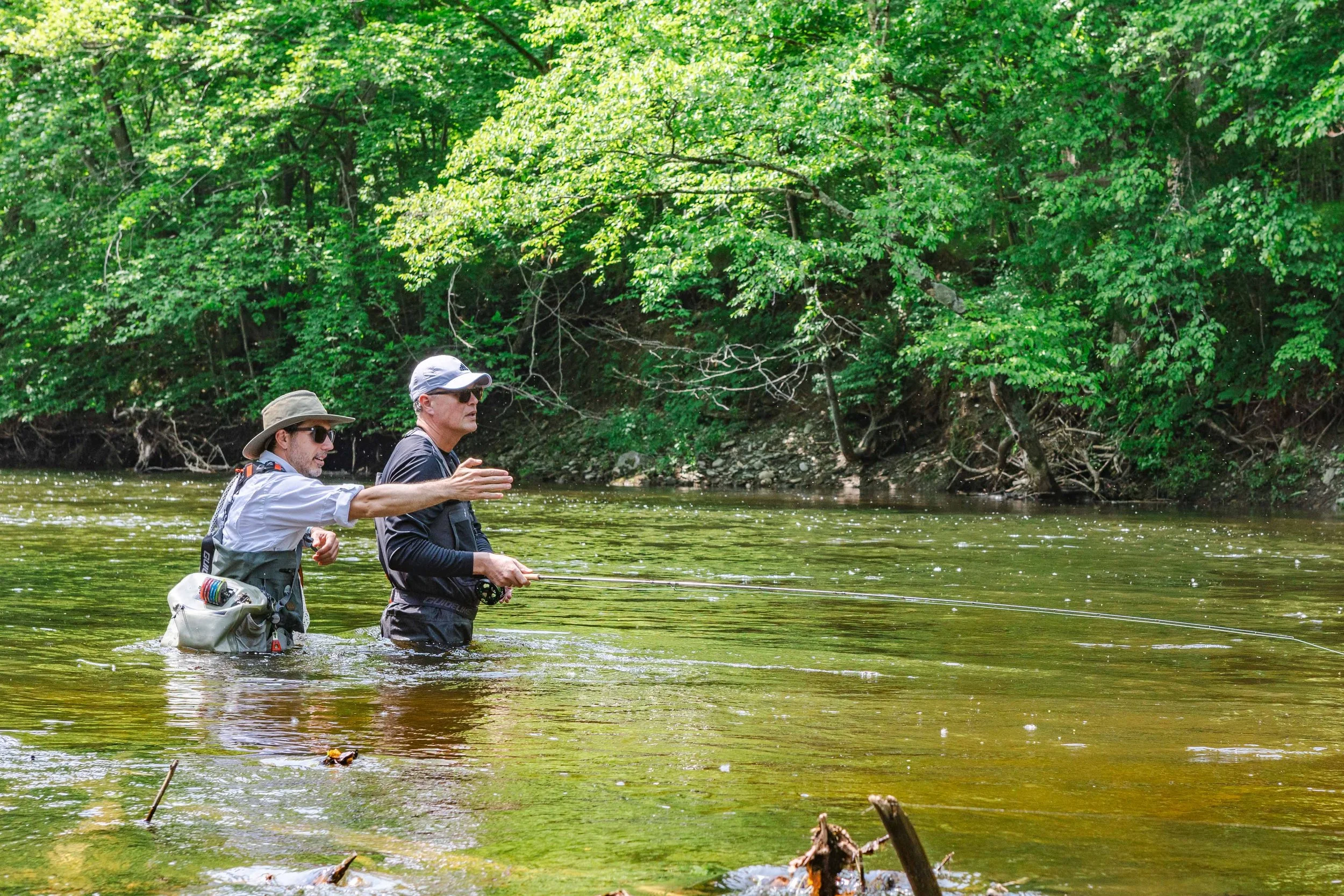 Two men fishing in a shallow river with lush green trees in the background.