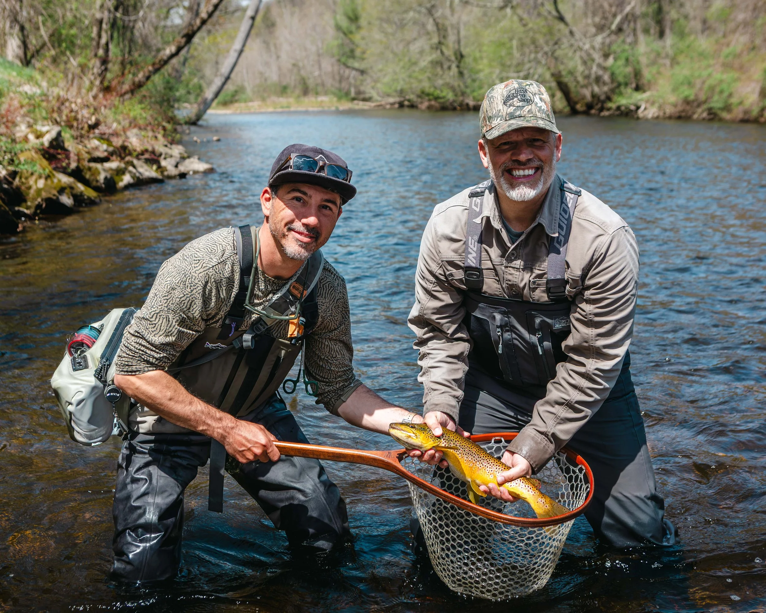 Two men standing in a river, holding a large rainbow trout with a fishing net in front of them, smiling at the camera.