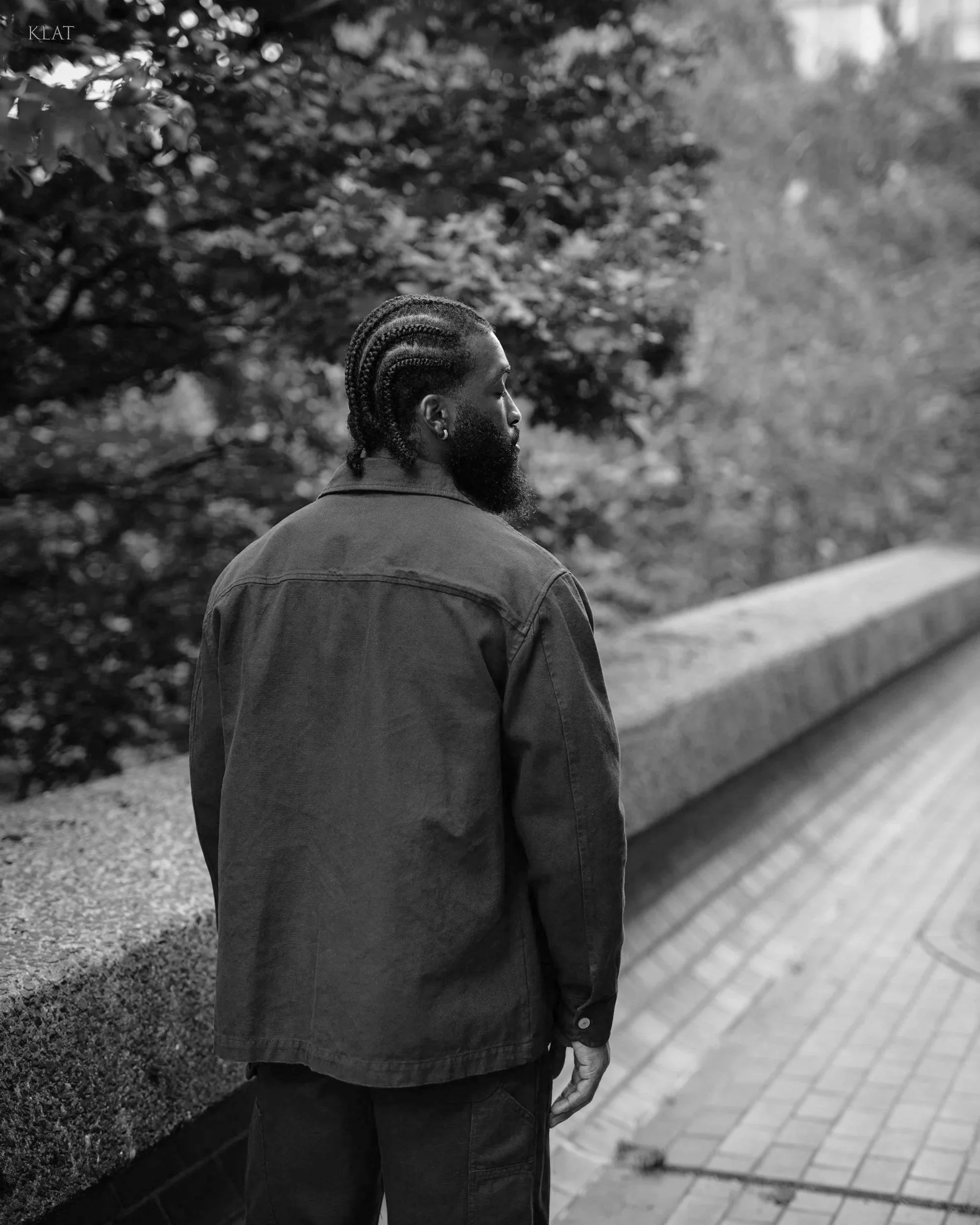 A man with braided hair and a beard standing near a stone wall, looking to the side, in a black and white photo.