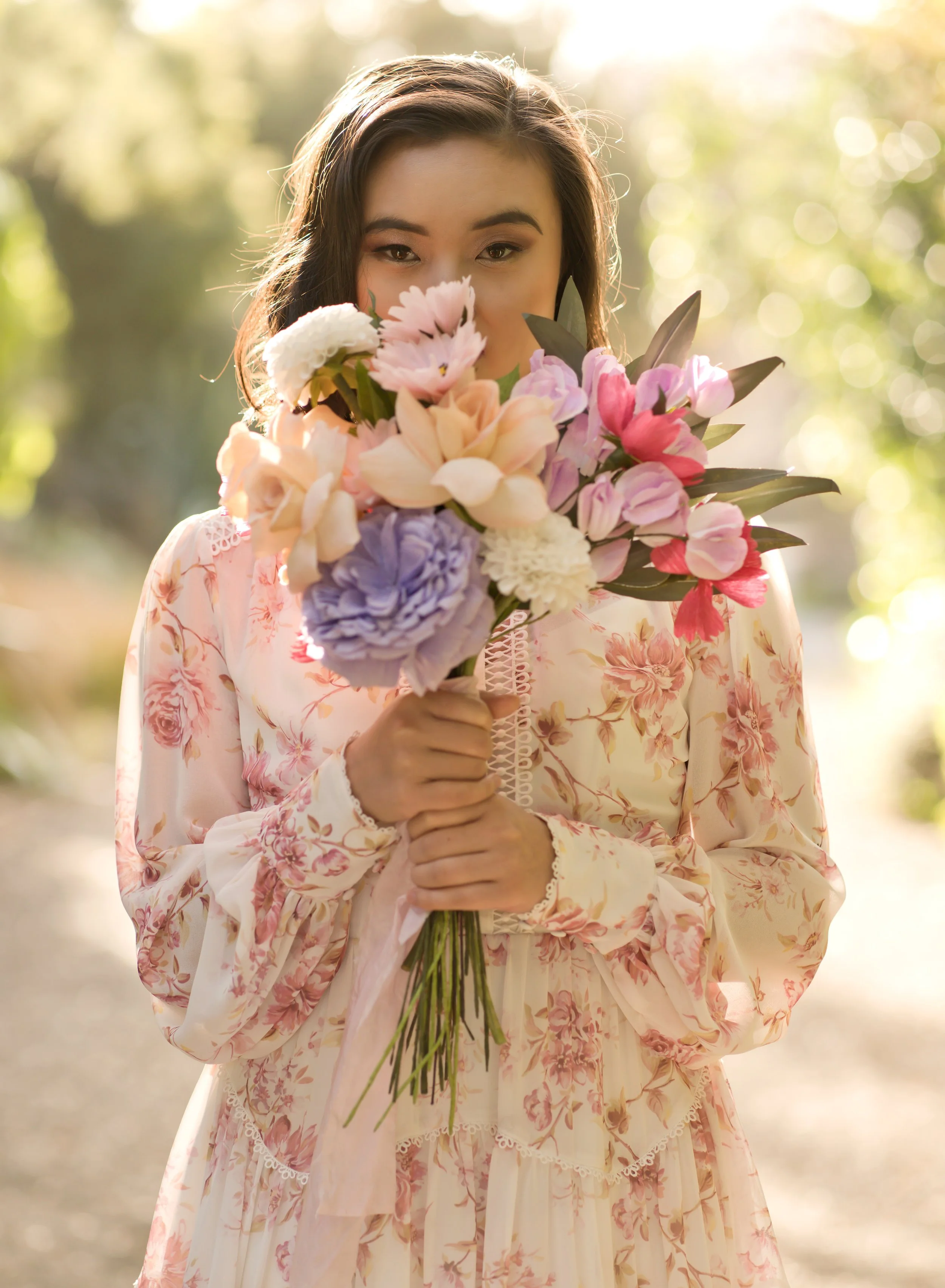A woman in a floral dress holding a bouquet of pink, purple, white, and beige flowers outdoors.