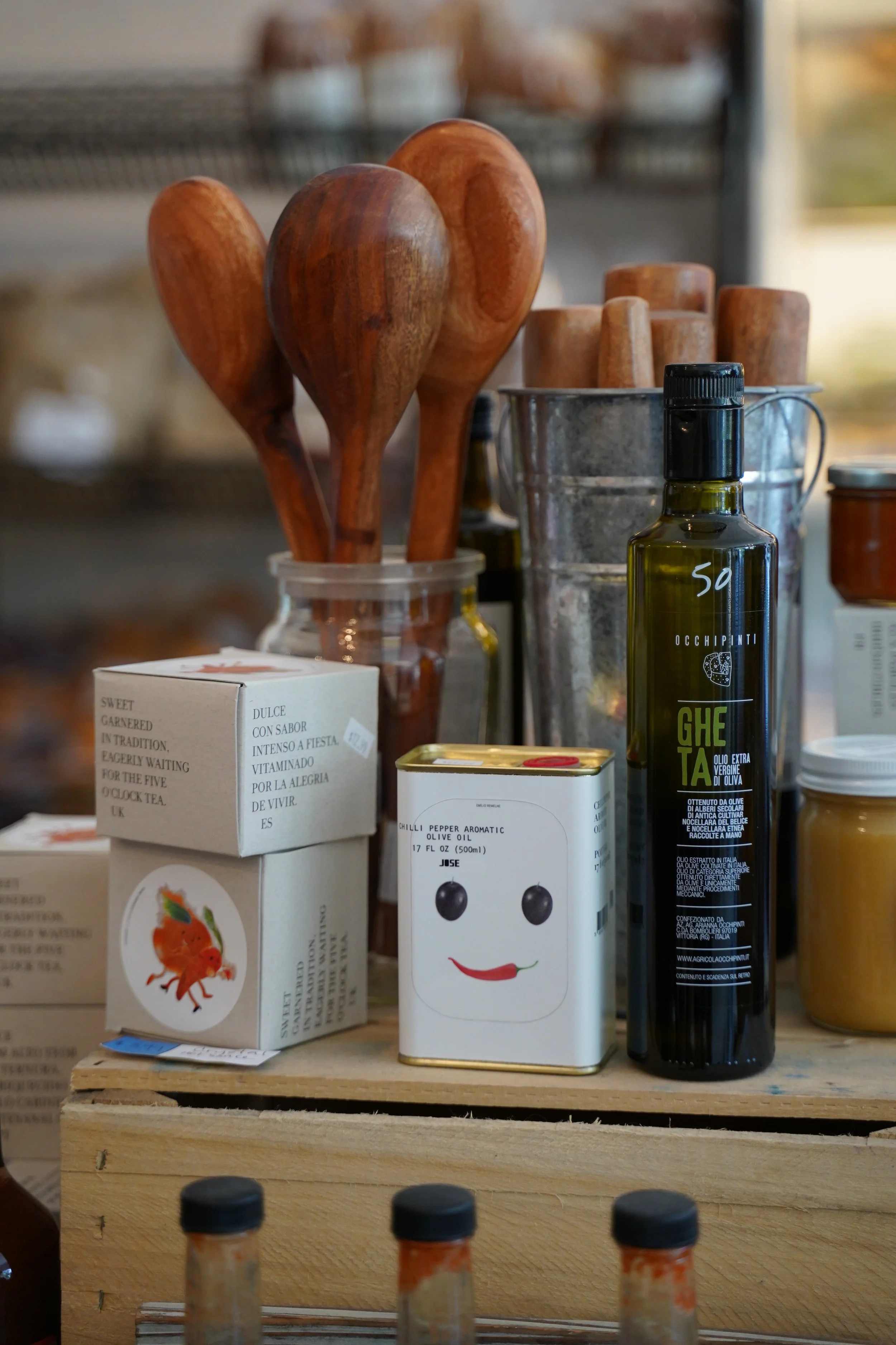 Assorted food products including a bottle of olive oil, boxes of seasonings, and wooden utensils on a wooden surface, available at Little House Green Grocery.