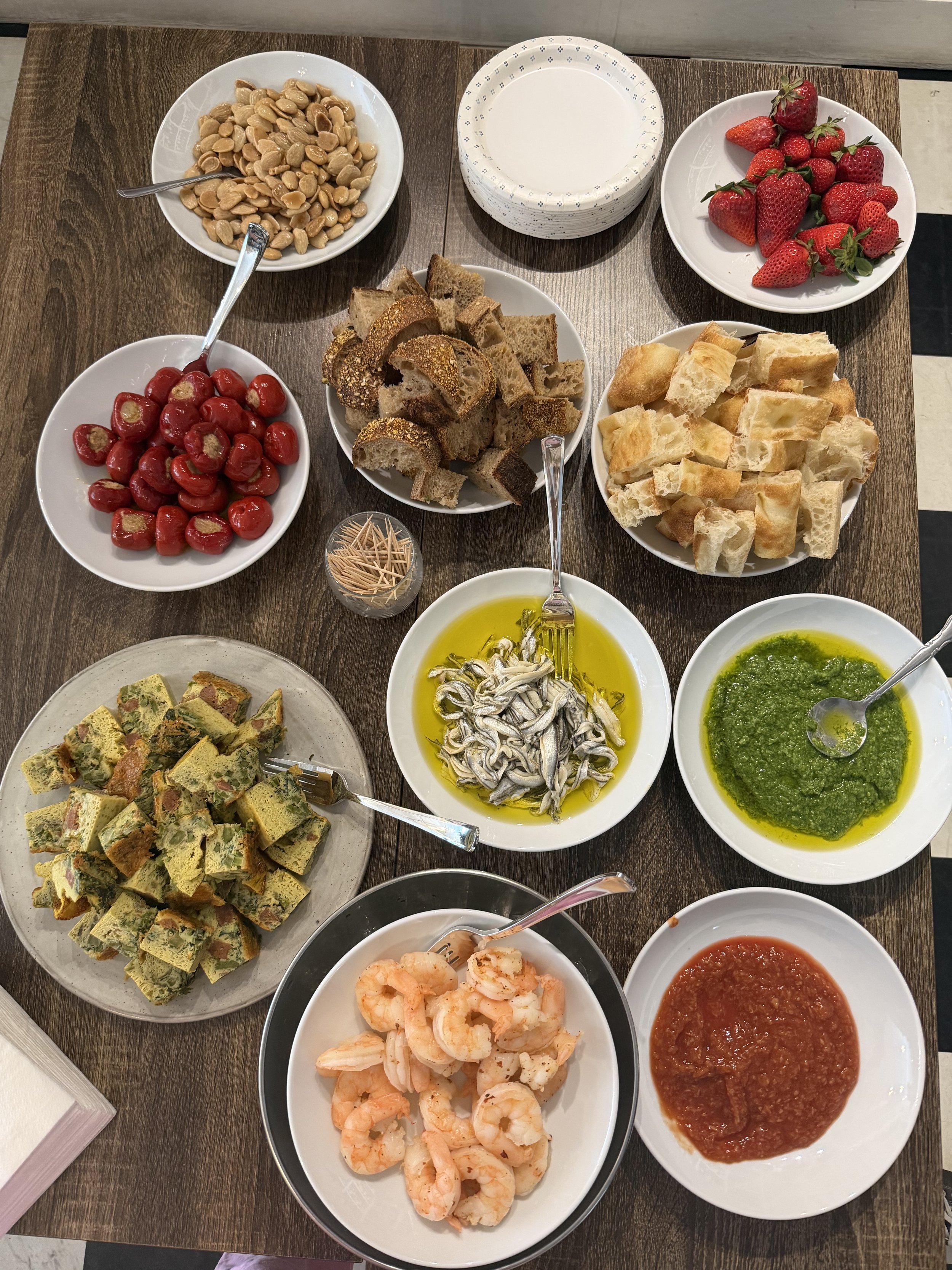 An assortment of dishes on a wooden table, including bowls of shrimp, bread, strawberries, cherries, various sauces, and small fish in olive oil. Options for catering from Little House Green Grocery.