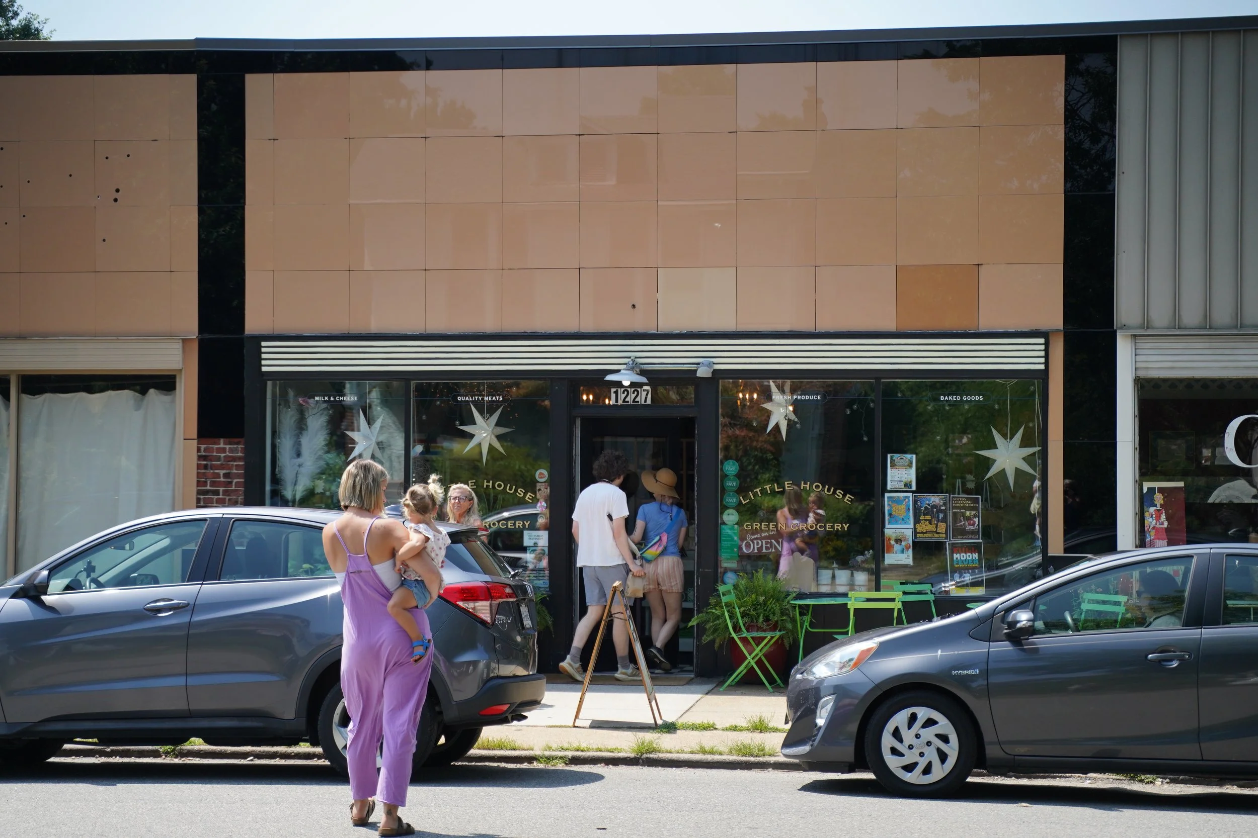 People standing and walking outside a grocery store called Little House Green Grocery in Bellevue neighborhood, with cars parked in front, on a sunny day.