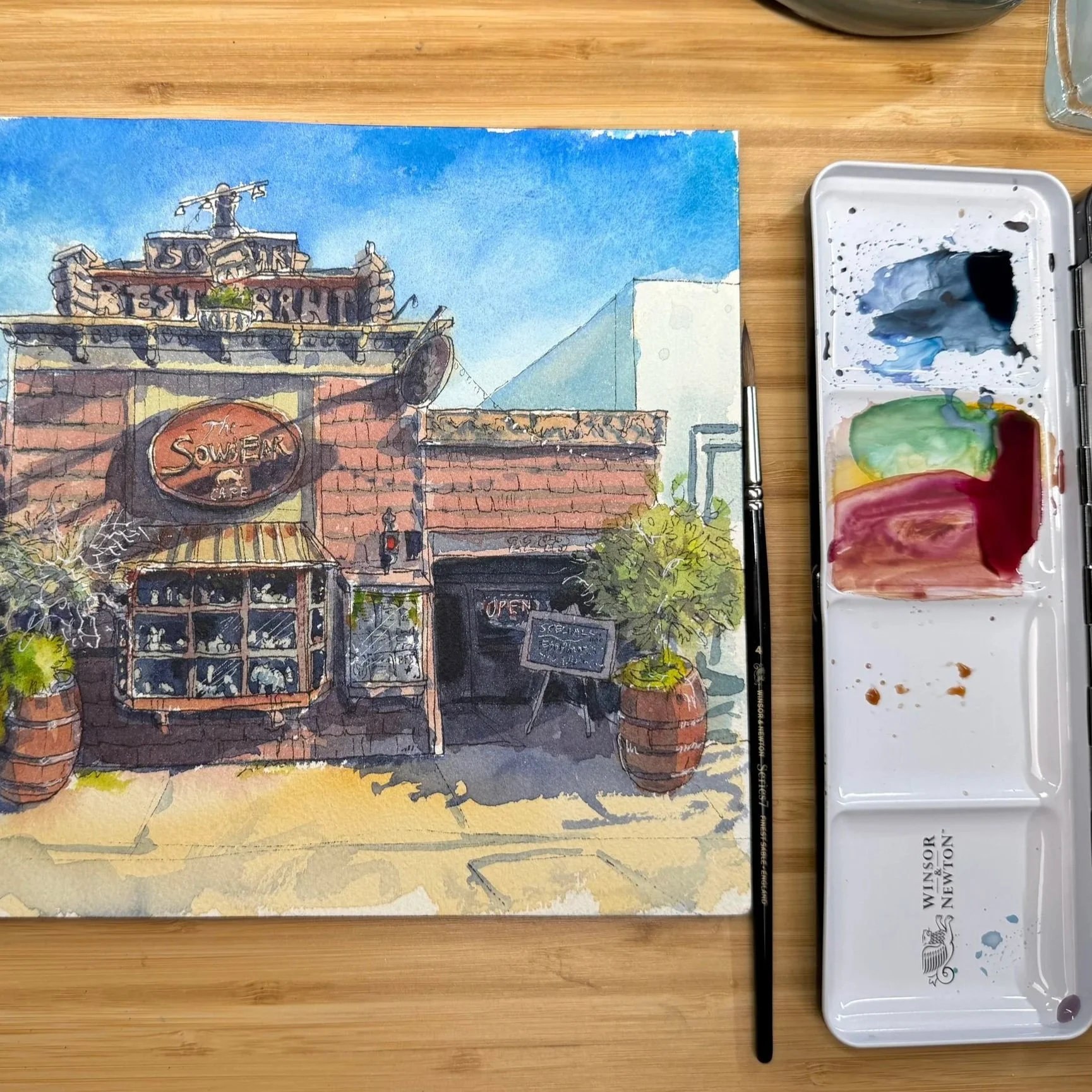 A watercolor painting of a restaurant facade with a brick and wood exterior, a sign reading "The Sower" and a rooftop sign that says "50th Year Restaurant"; the painting is on a wooden surface next to a watercolor palette with various colors and a black brush.