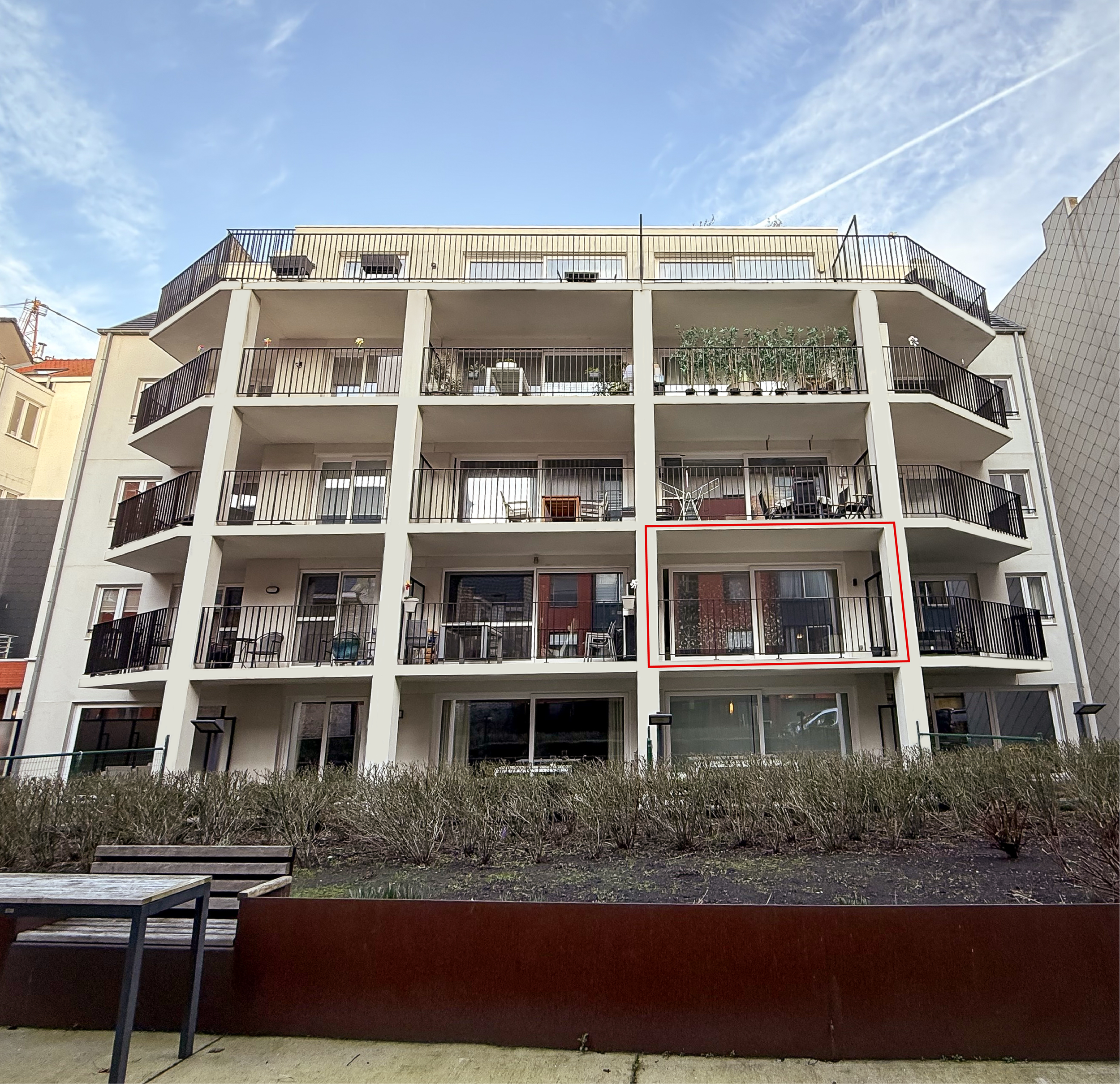 Bovenaanzicht van een wit appartementencomplex met meerdere verdiepingen en balkons, met een blauwe lucht en wolken op de achtergrond, en een tuin met bankjes op de voorgrond, vertigo oostende, vertigo, appartement oostende, appartement te koop