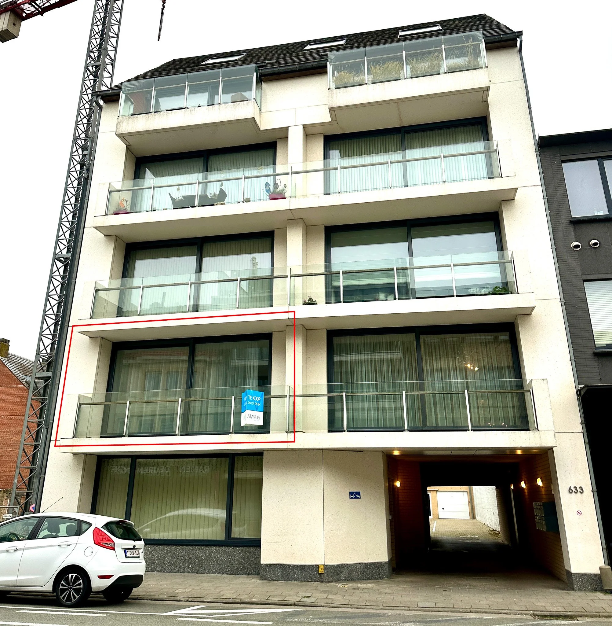 Vier verdiepingen hoog modern appartementencomplex met glazen balustrades, een onderbouw met een garage en een inrit, een auto geparkeerd voor het gebouw, en een teken dat de woning te koop is.