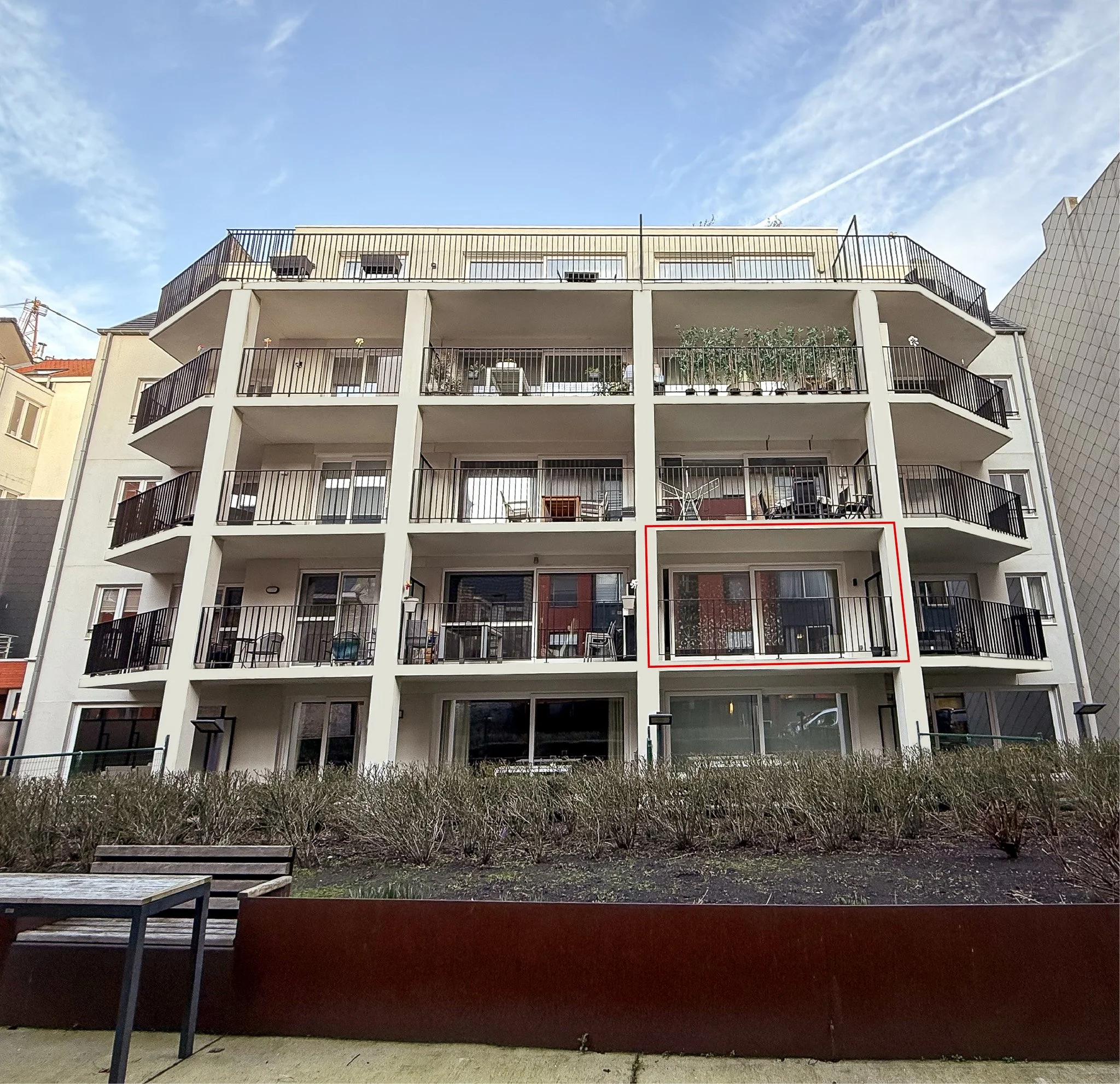 Een wit appartementencomplex met vier verdiepingen, elke verdieping heeft een balkon met zwarte railing. Het balkon op de derde verdieping is gemarkeerd met een rood kader. Het gebouw staat voor een tuin met struiken en een bank, vertigo oostende