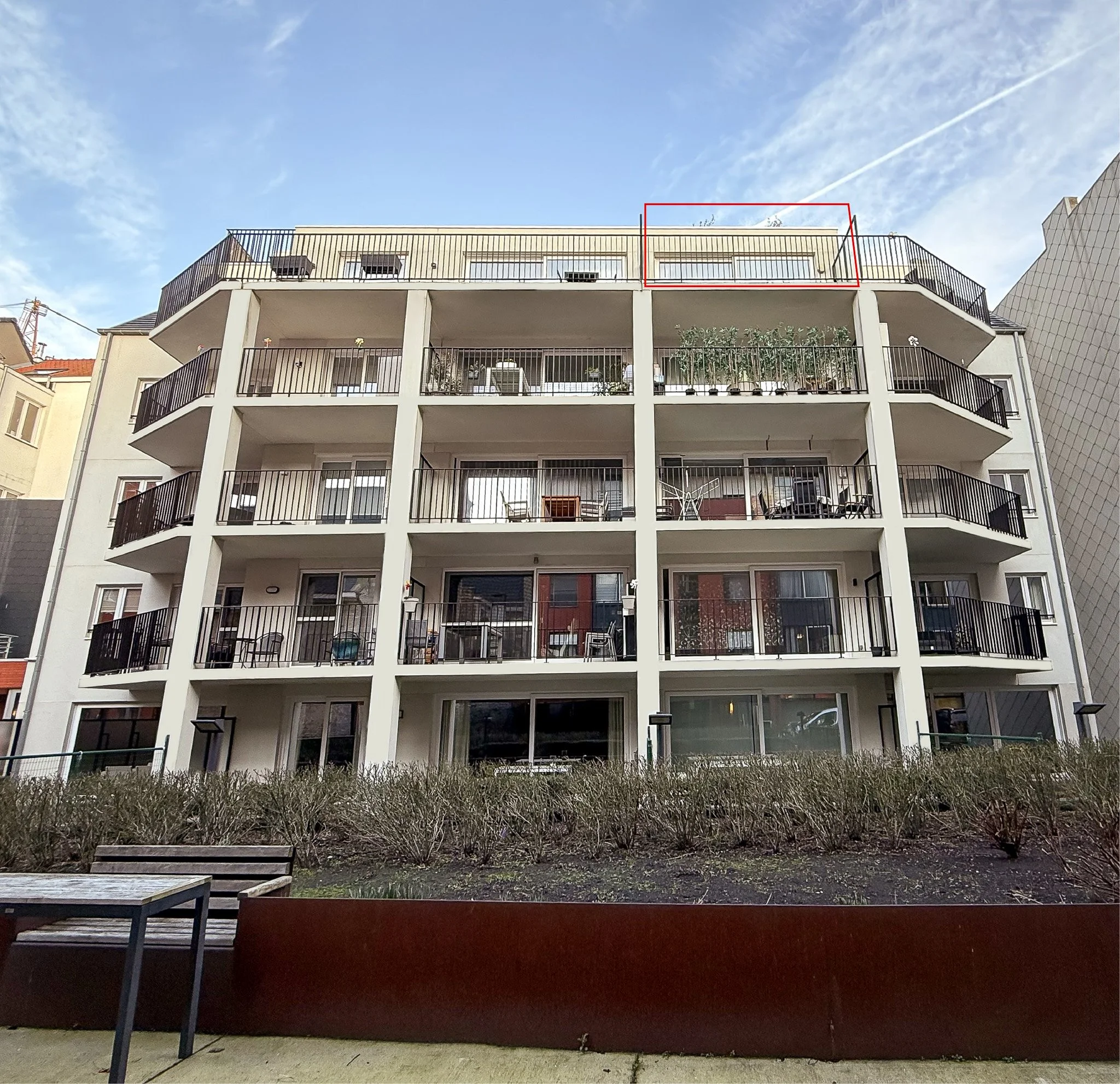 Een wit vier verdiepingen tellend appartementencomplex met balkons en een tuin op de begane grond, onder een blauwe hemel, appartement vertigo te koop, appartement oostende te koop