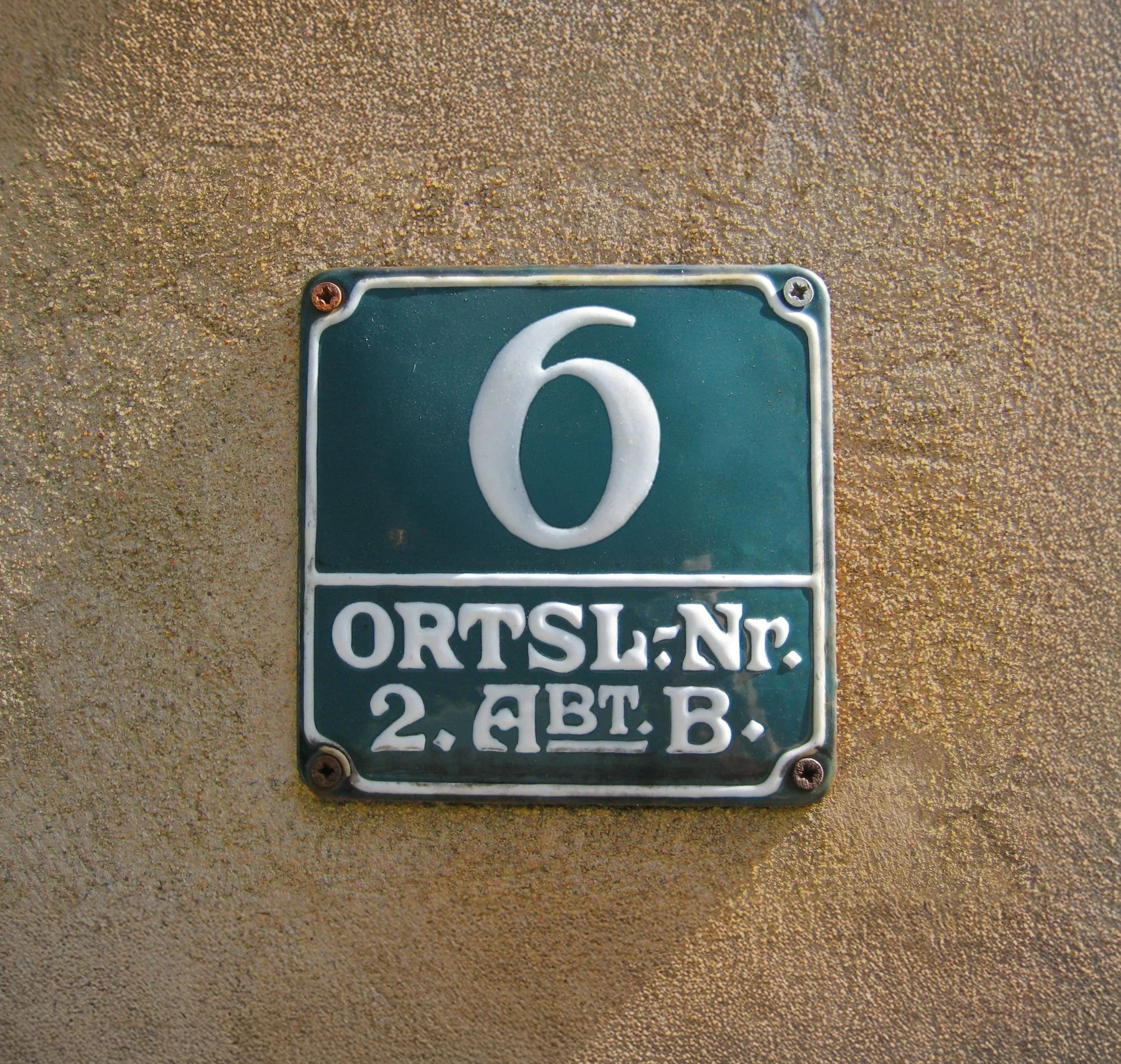 Ein metallener Straßenschild mit der Nummer 6, befestigt an einer sandigen Wand.