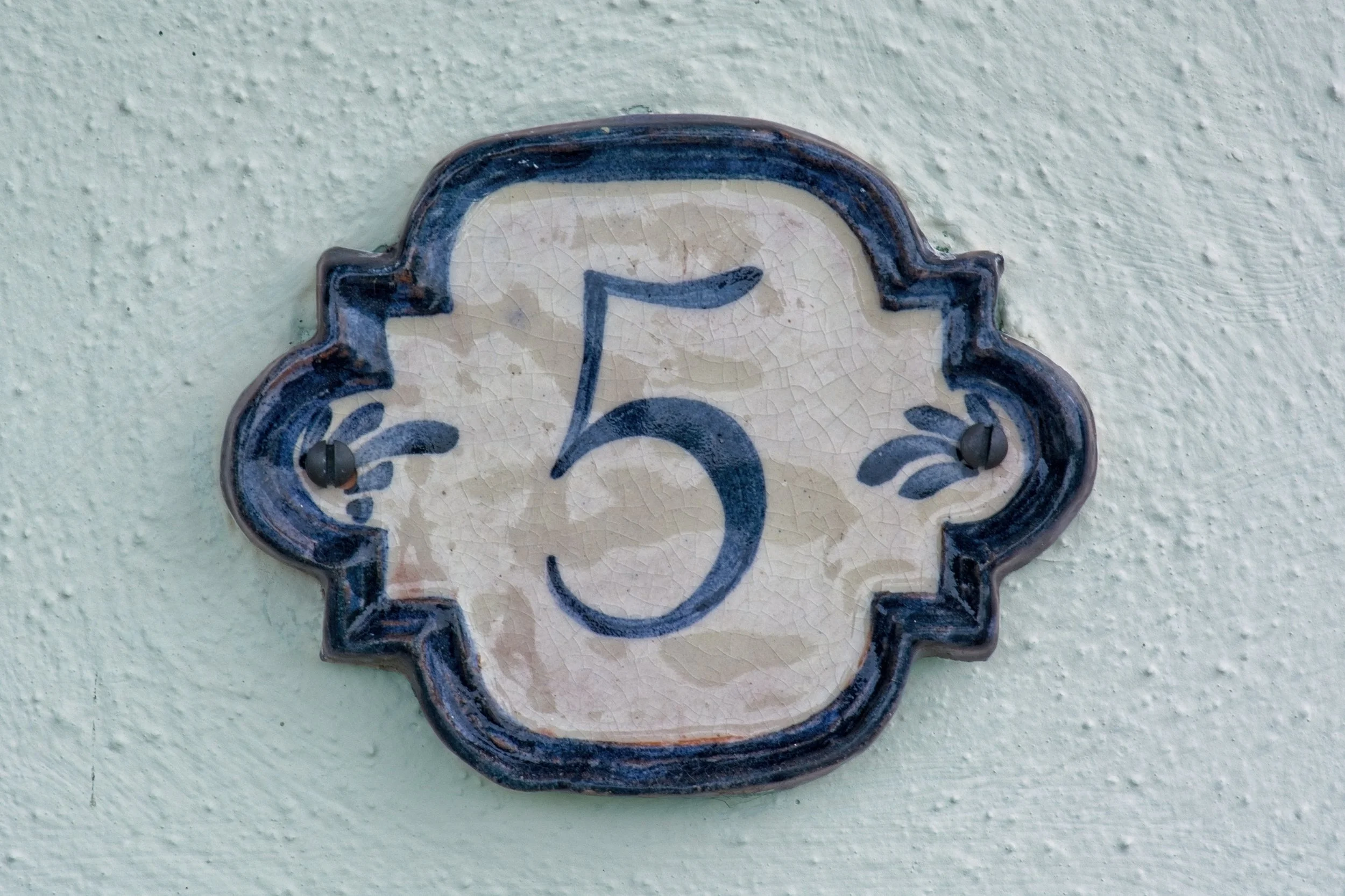 Keramikplakette mit der Hausnummer 5, befestigt an einer hellgrünen Wand.