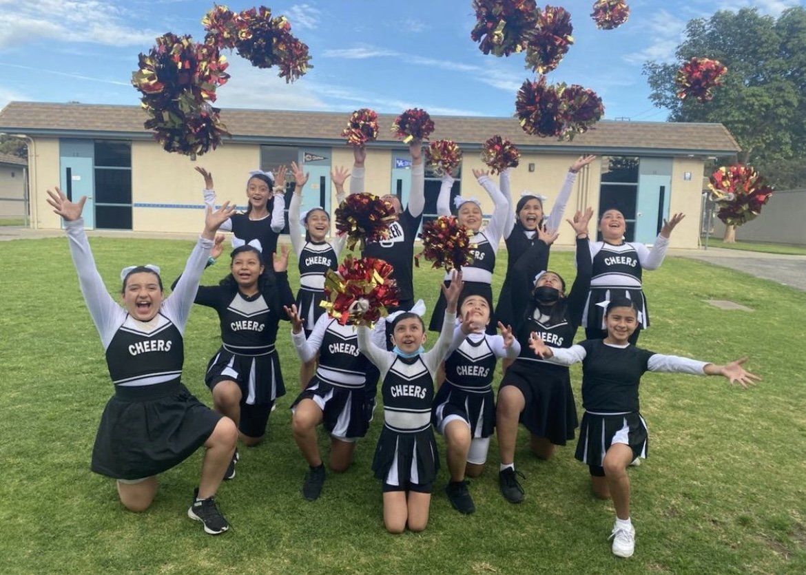 Group of young cheerleaders in black and white uniforms celebrating outdoors, throwing pom-poms in the air, smiling, and posing on a grassy field with a building in the background.