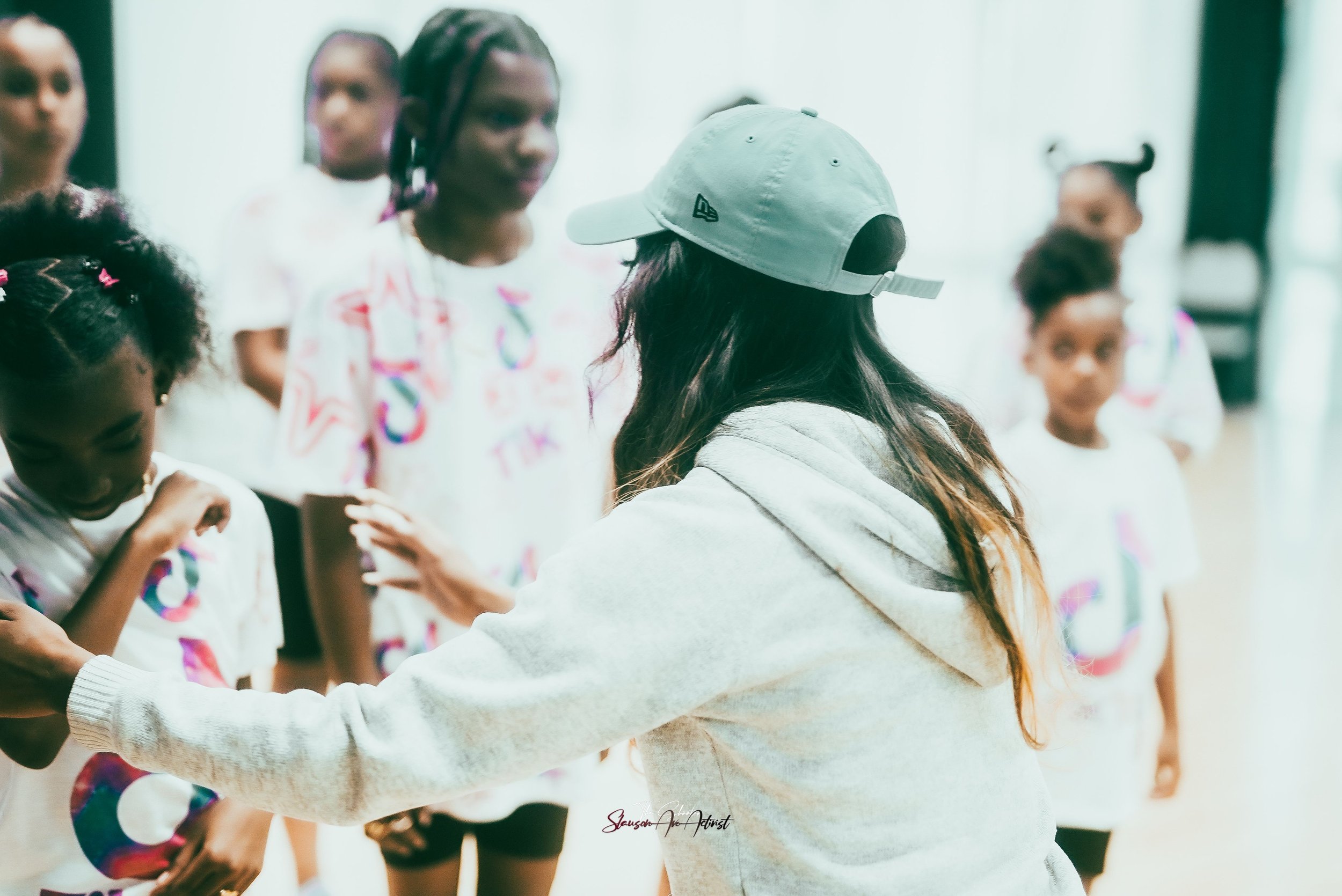 A group of children in white graphic T-shirts standing in a line with a woman wearing a light blue baseball cap and gray hoodie directing them.