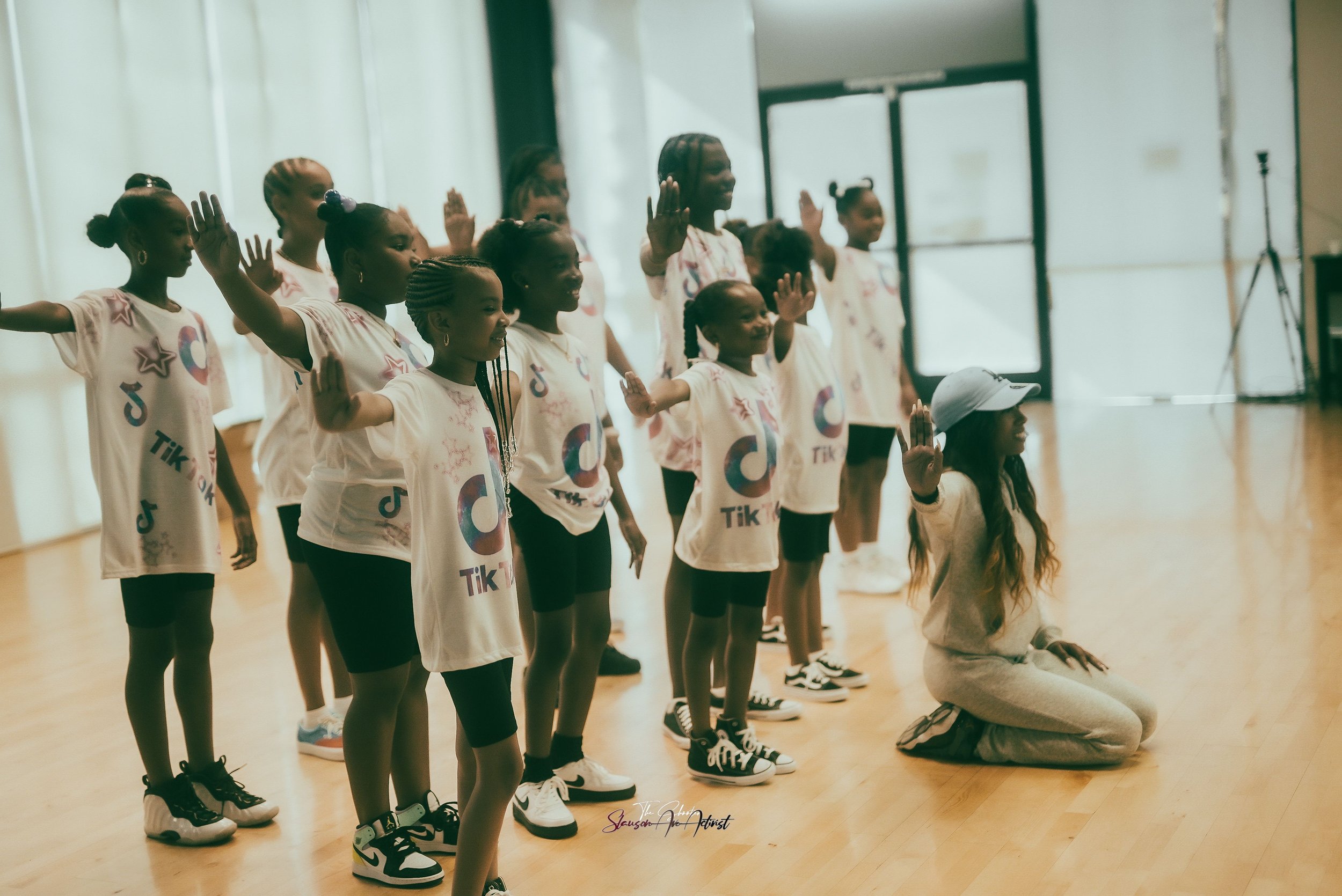 A group of young girls and a woman practicing dance moves in a dance studio, all wearing matching white T-shirts with TikTok logos, some with raised hands.