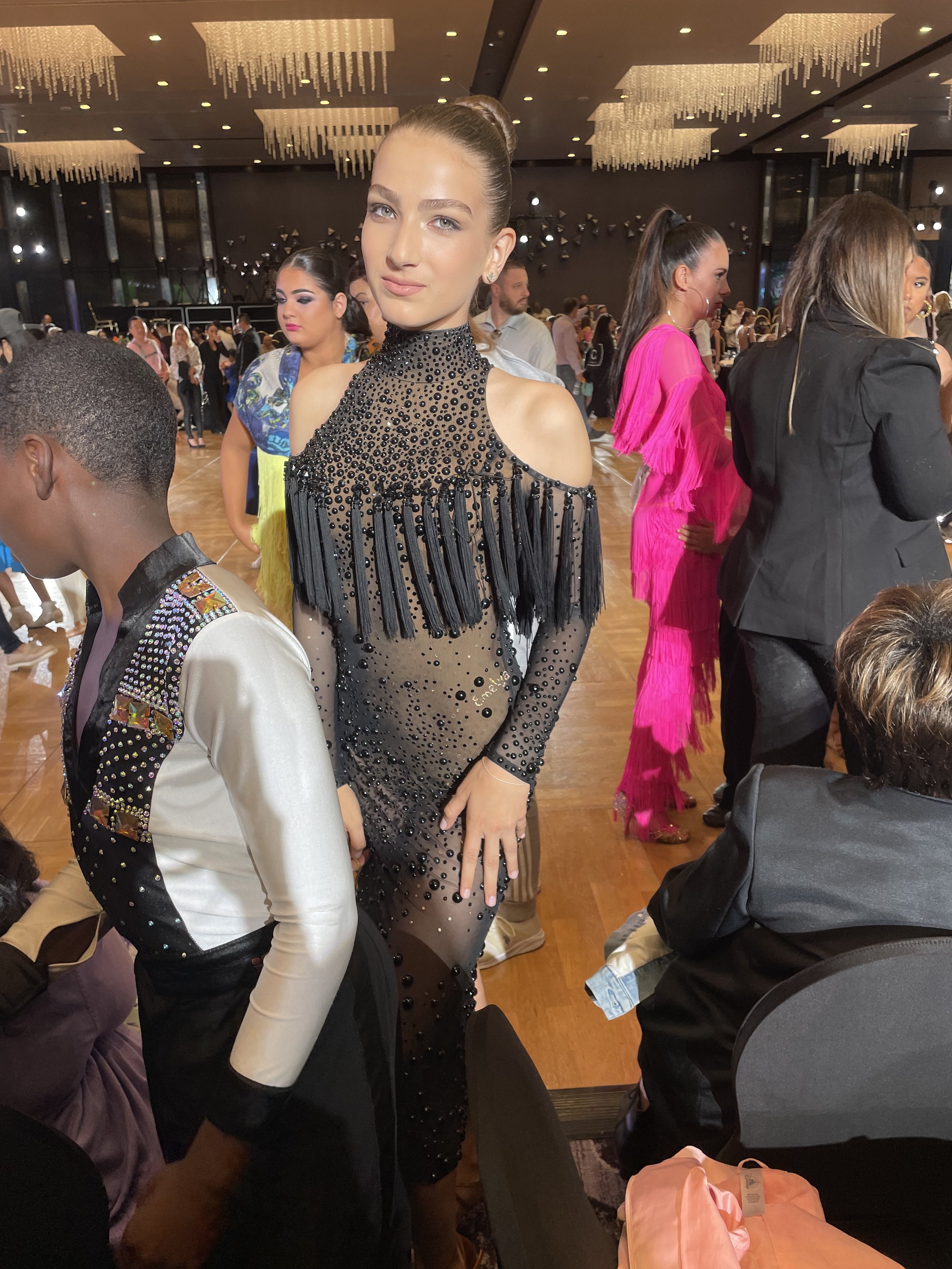 A fashionable event - ballroom competition - featuring a young dancer in a black fringed dress, surrounded by people wearing colorful clothing, in a large, well-lit room with chandeliers.
