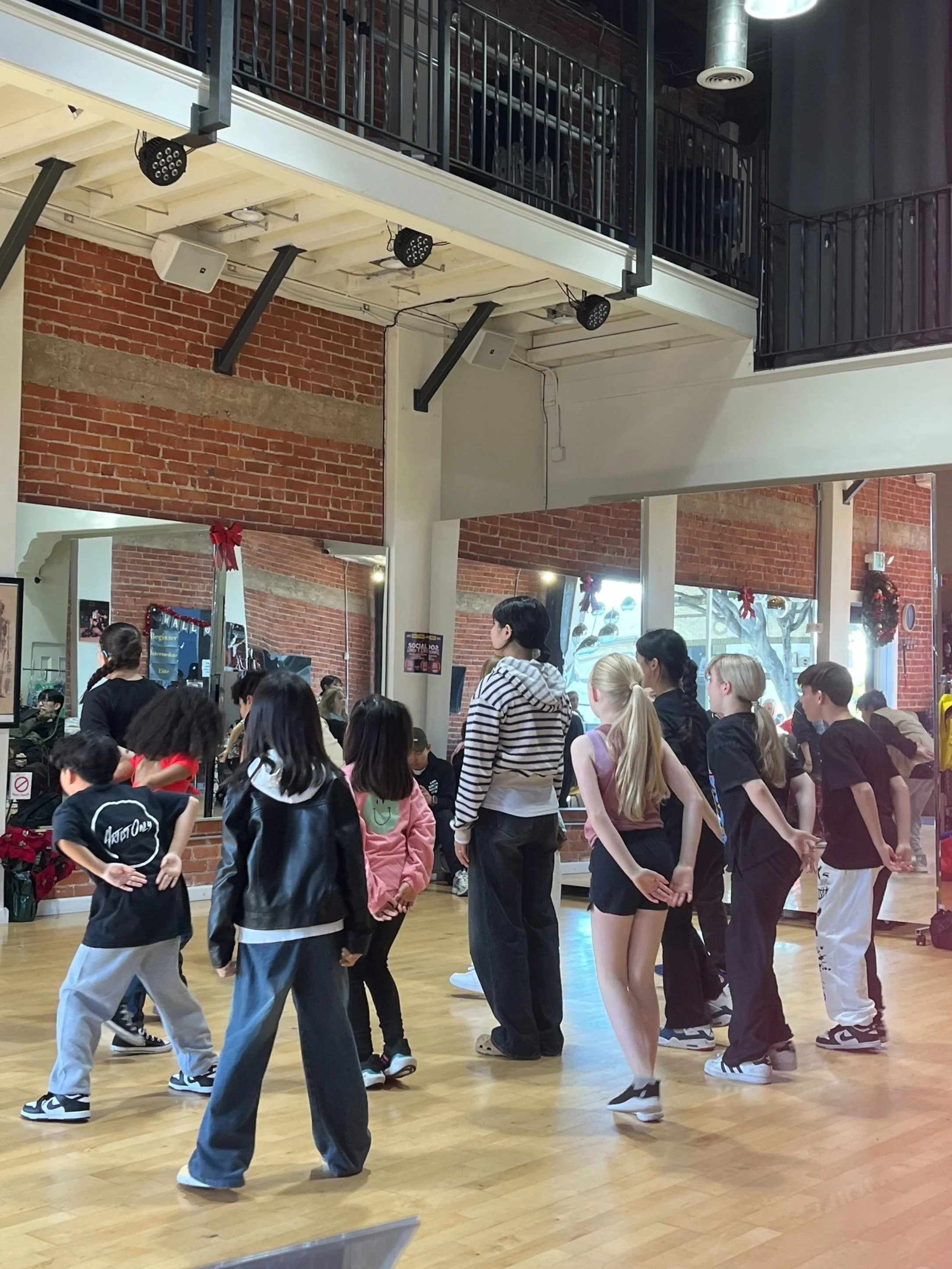 A group of children dancin in a dance studio with wooden floors, brick walls, and mirrors. They appear to be in a dance class, wearing casual clothes, with some children facing forward and others turned away. The ceiling has exposed beams and lights.