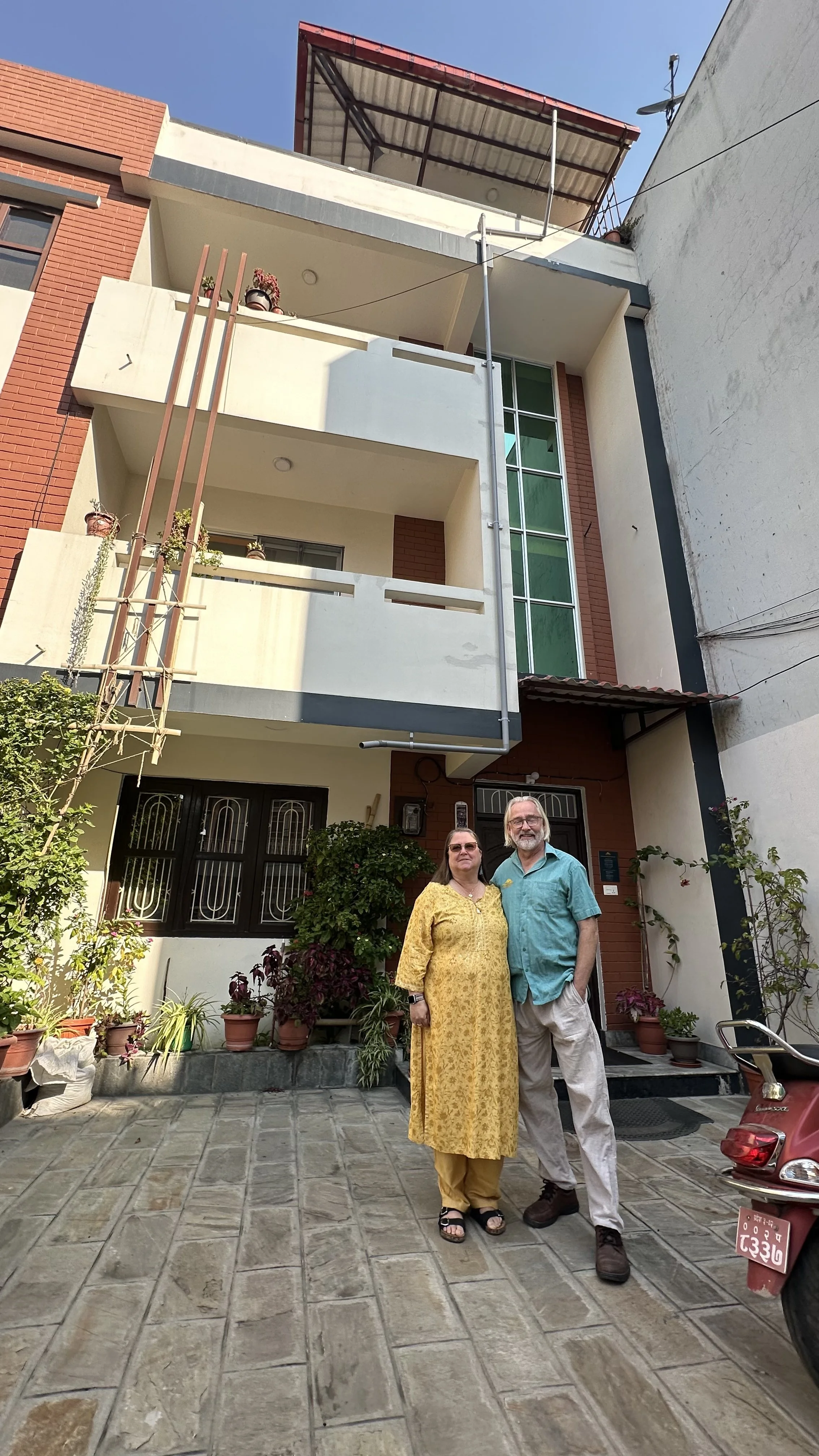A couple standing in front of a multi-story house with potted plants and a garden. The woman is wearing a yellow traditional dress, and the man is wearing a turquoise shirt and khaki pants.