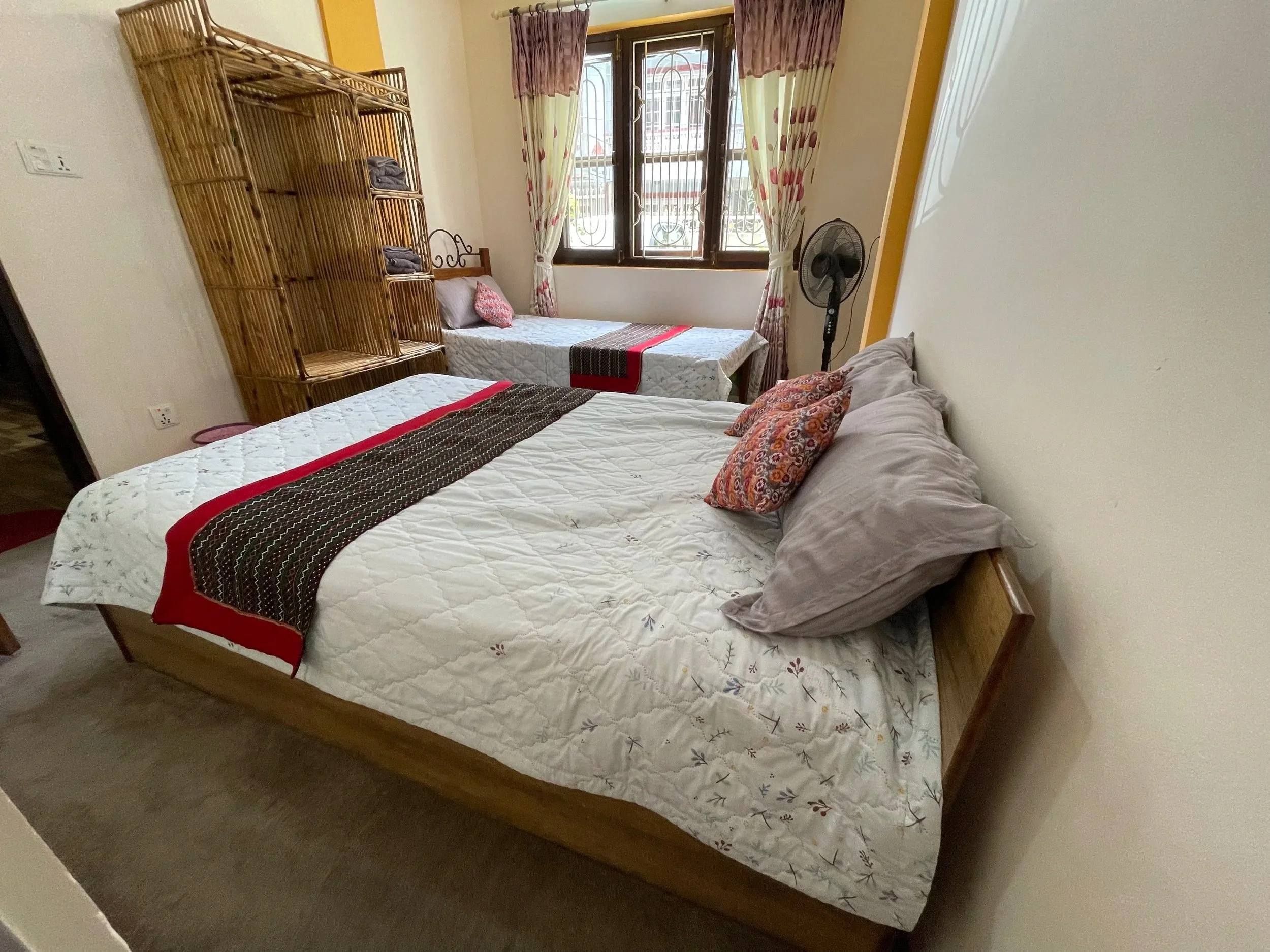 A bedroom with a bed in the foreground, covered with a white quilt that has floral designs, and a decorative pillow. There are two beds, one in the background near a window with pink and cream curtains, and another in the foreground with colorful pillows. A wooden headboard is visible, and there is a standing fan next to the window. The room has beige walls and a window with a view of buildings outside.