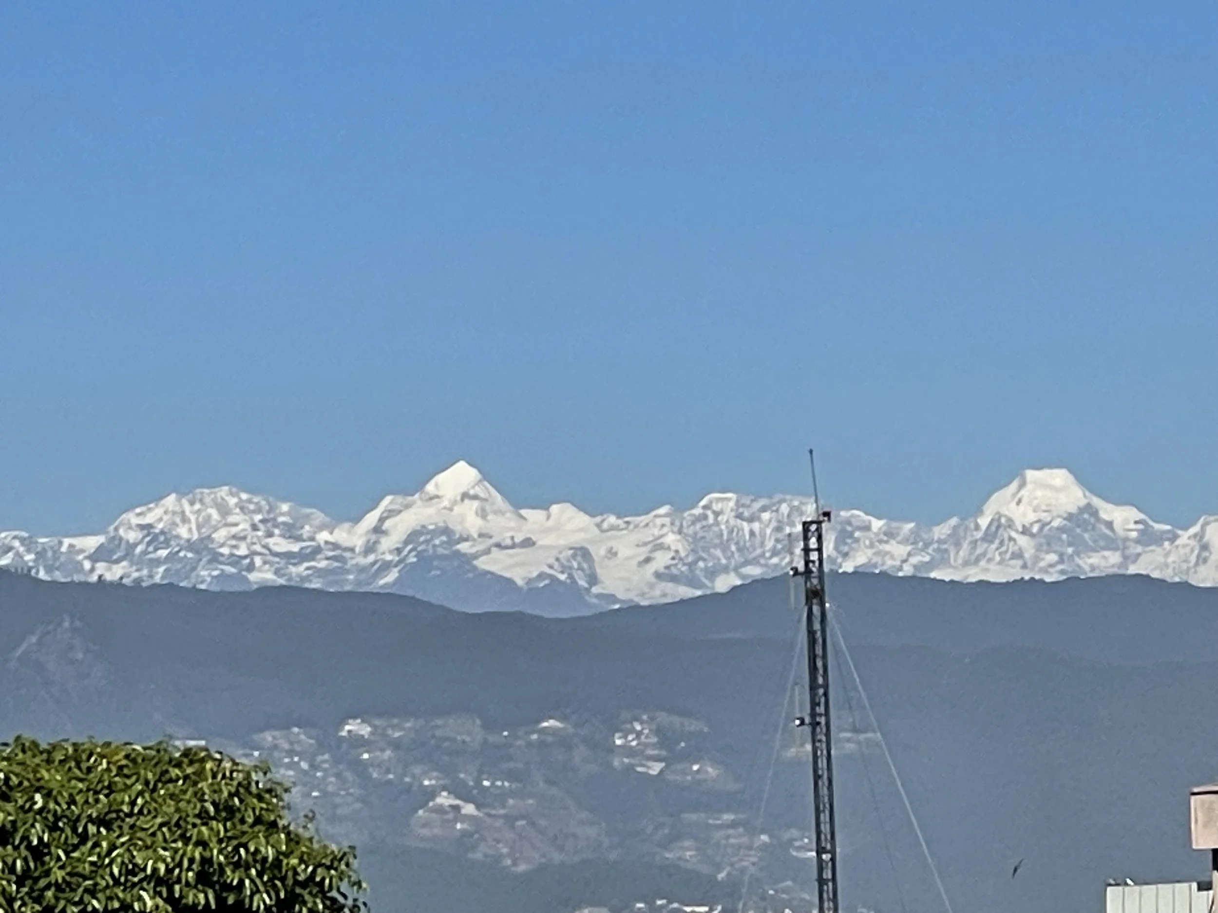 Snow-capped Himalayan mountains under a clear blue sky with a tall communication tower in the foreground.